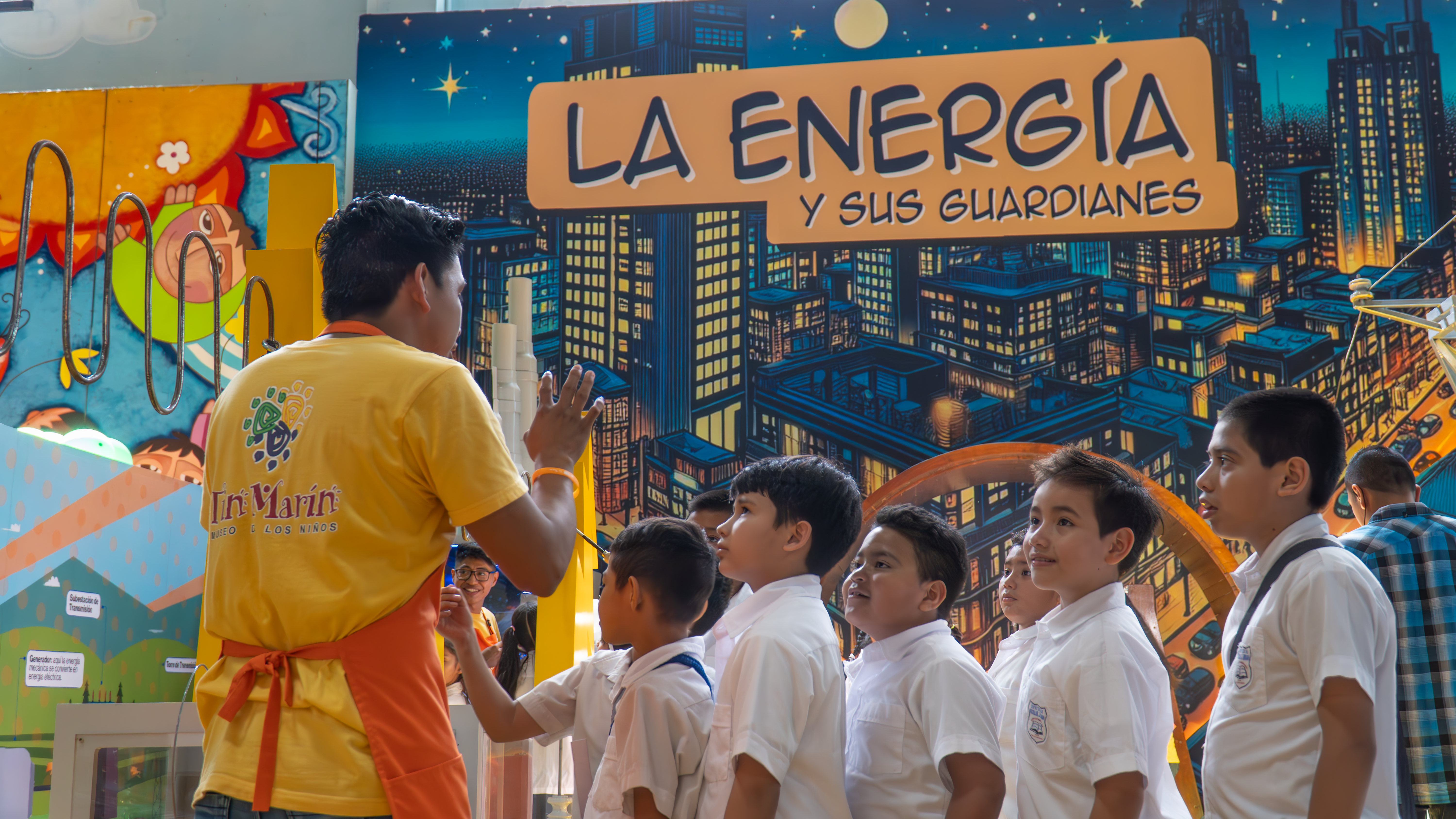 group of young students in white uniforms attentively listen to a staff member wearing a yellow Tin Marín apron as he explains an interactive energy exhibit. The background features a vibrant cityscape illustration with the words "La Energía y sus Guardianes." The exhibit showcases educational elements related to electricity and sustainability in an engaging museum setting.