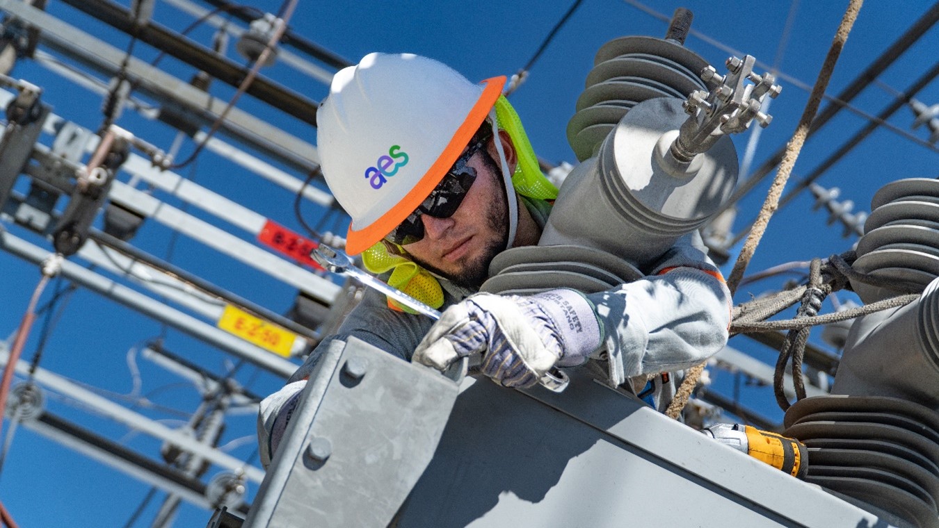 A lineman wearing a white hard hat with "aes" in blue and orange, safety glasses, and gloves works on electrical equipment. He is positioned high up amidst a network of power lines, insulators, and transformers against a clear blue sky. He appears to be securing or adjusting a connection.