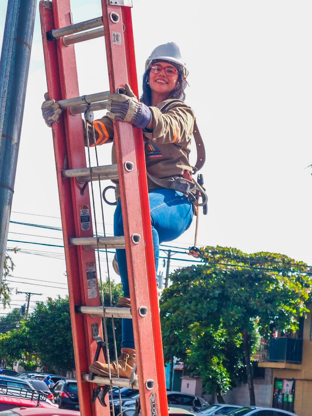 Una mujer sonriente con gafas, un casco blanco y guantes de trabajo está subiendo una escalera roja. Lleva una chaqueta marrón, pantalones azules y botas marrones. La escalera parece estar apoyada contra un poste vertical. En el fondo se ven árboles, coches aparcados y un cielo brillante.