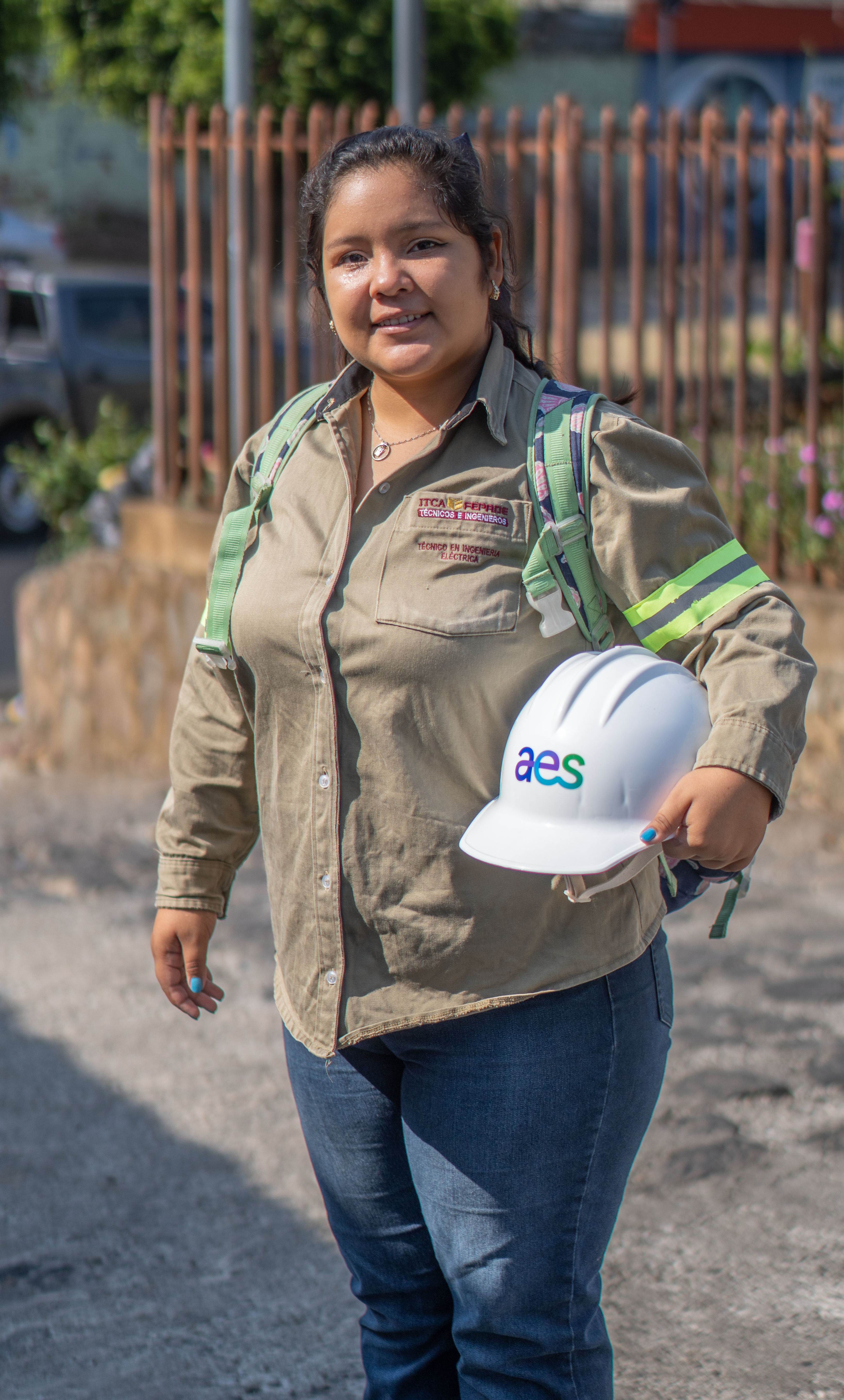A young woman wearing a khaki shirt with an embroidered electrical engineering program logo and reflective armband stands outdoors holding a white AES hard hat. She has a green and purple backpack on and is smiling at the camera with a sunny, urban background featuring a fence and greenery.