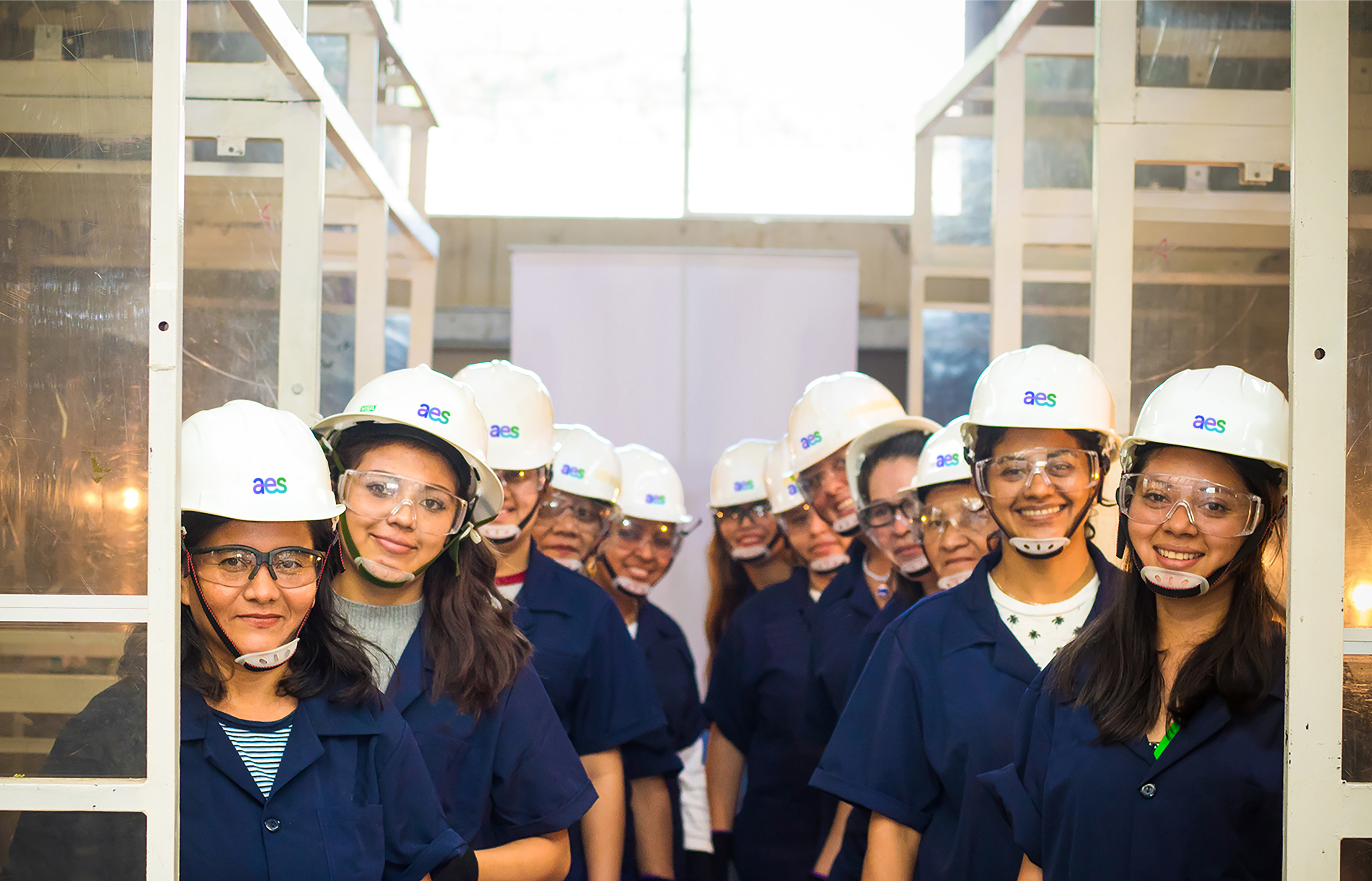 A group of approximately ten students wearing white hard hats with the letters "aes" in blue and clear safety goggles smile and look towards the camera. They are wearing dark blue jackets or shirts. The setting appears to be an industrial or educational environment with metal framing visible around them and a white wall in the background.