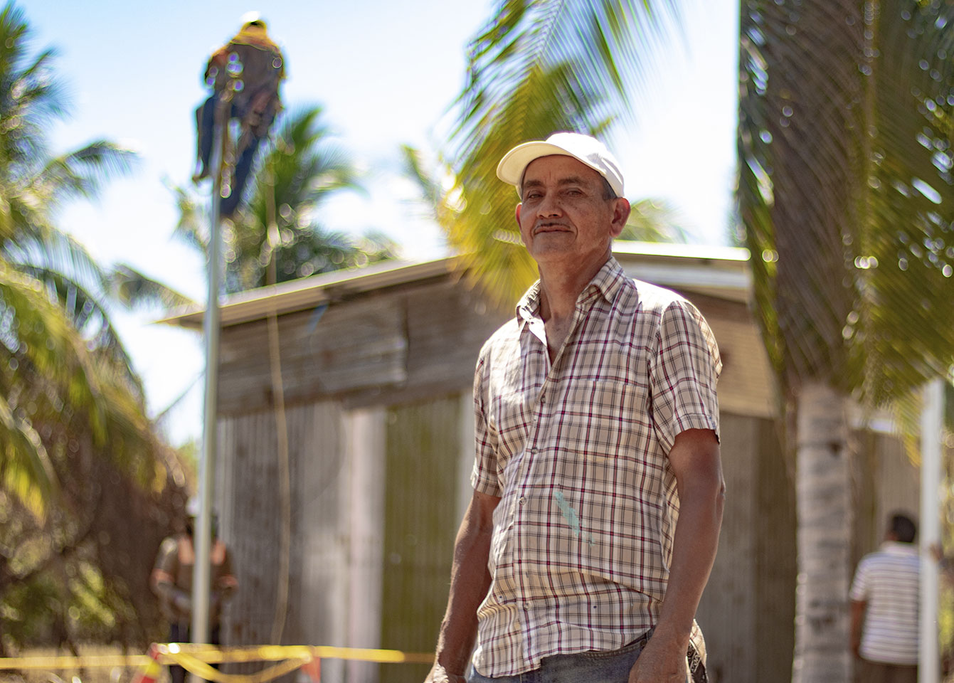 An older man in a plaid shirt and white cap stands in front of a rural home surrounded by palm trees. In the background, an electrical worker is high on a pole performing maintenance or installation, while another worker stands below. The scene is bright and sunny, with yellow caution tape visible on the ground.