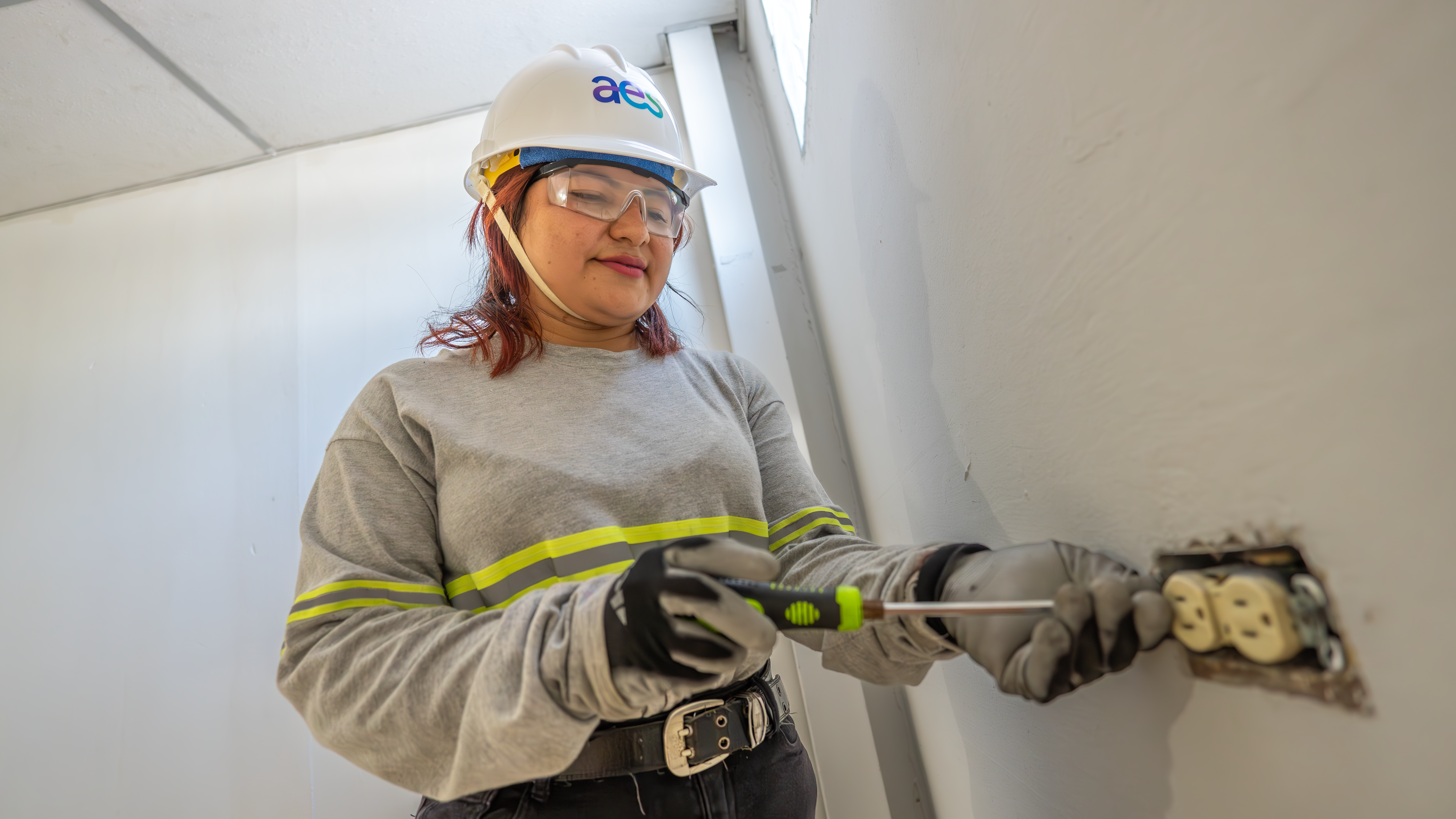 mujer-electricista-trabajando-en-toma-de-corriente Una mujer con casco de seguridad blanco, gafas protectoras y guantes de trabajo está ajustando un enchufe eléctrico de pared con un destornillador. Lleva una camisa gris de manga larga con rayas reflectantes amarillas. La pared es de color claro y se ven partes de la instalación eléctrica.