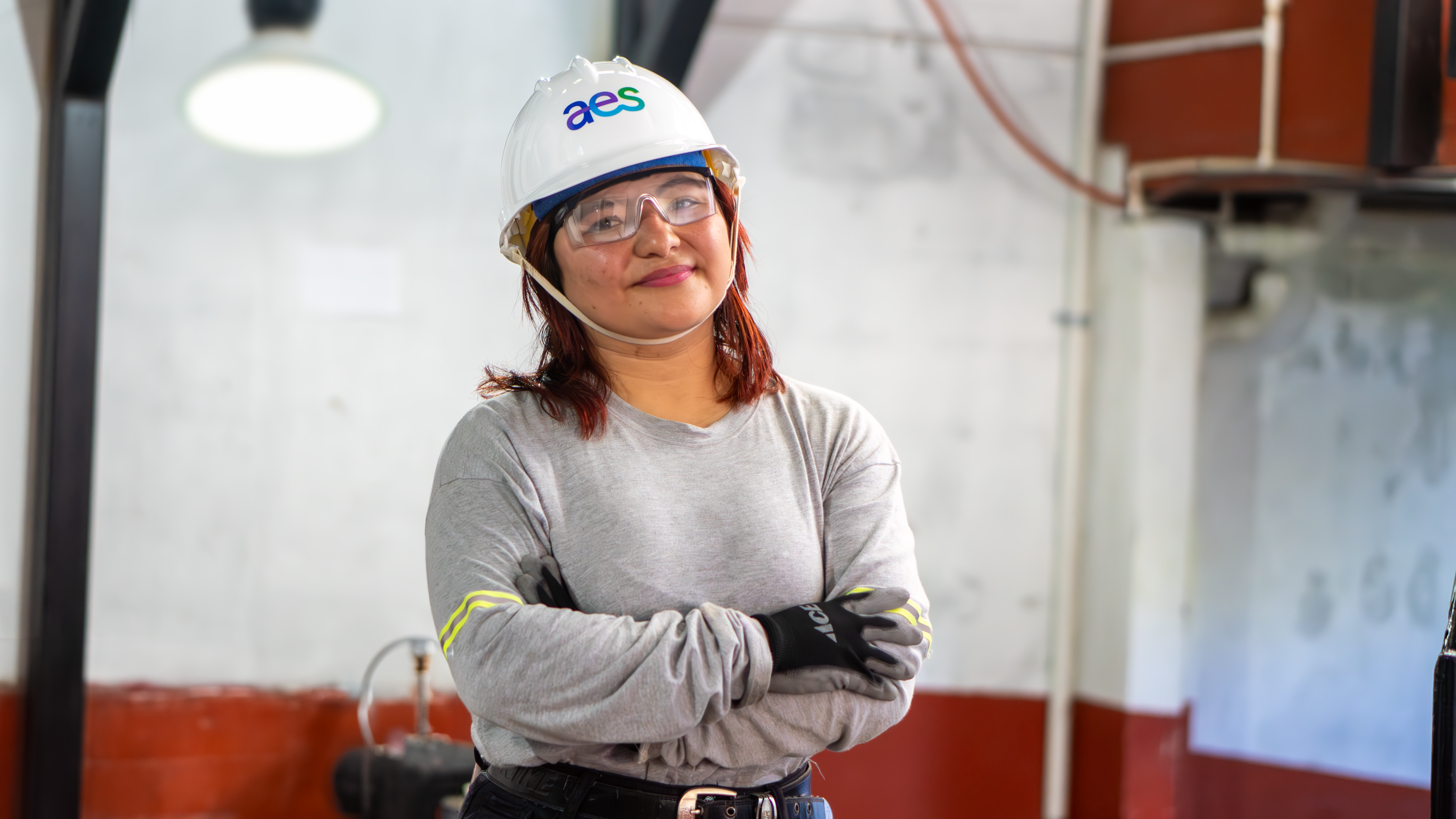 Una mujer sonríe a la cámara mientras está de pie con los brazos cruzados. Lleva un casco de seguridad blanco con el logo de "aes", gafas de seguridad transparentes, una camisa gris de manga larga con detalles amarillos en los puños y guantes de trabajo negros. Está en un entorno industrial con tuberías y equipos visibles al fondo.