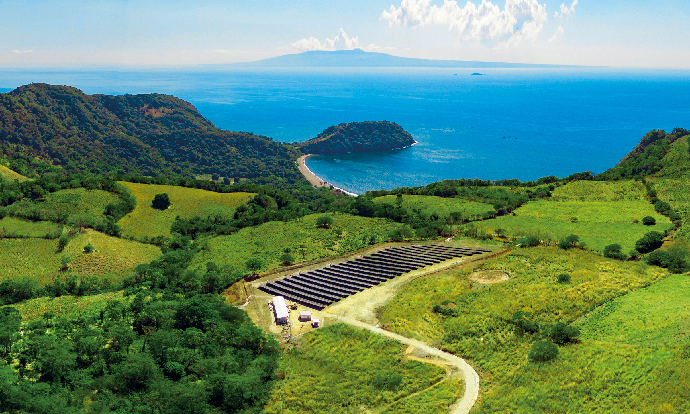 Aerial view of a large solar panel farm situated in a green, hilly landscape near a coastline with a sandy beach and blue ocean. Mountains are visible in the distance under a partly cloudy sky.