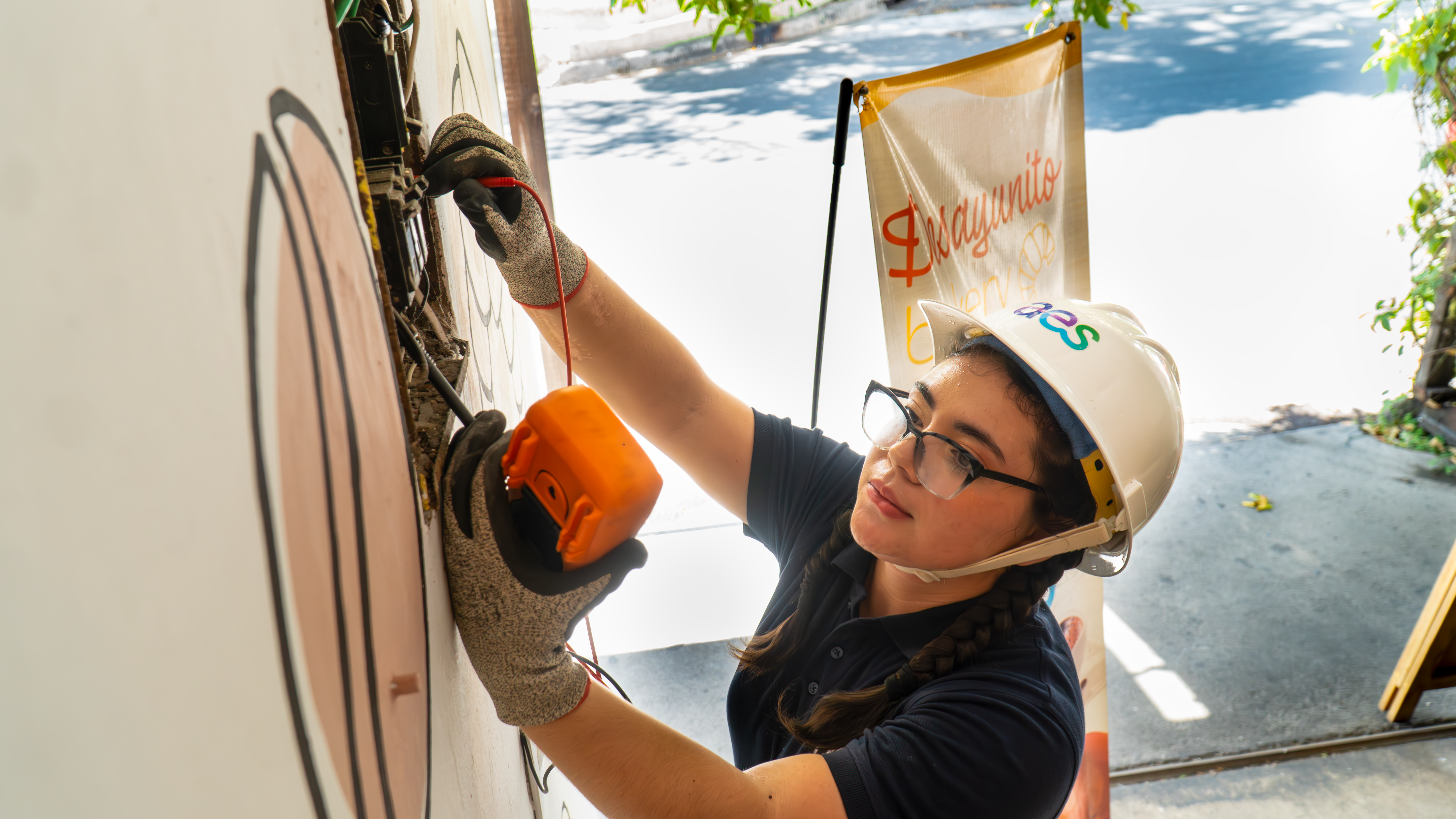 A young woman wearing an AES-branded white hard hat, safety glasses, and gloves is using an electrical tester to check a connection on a wall-mounted panel. She is focused on her task, holding the tester with one hand while securing the wires with the other. The background features a partially visible banner with the words "Desayunito" and "bueno" in an outdoor setting with sunlight filtering through trees.