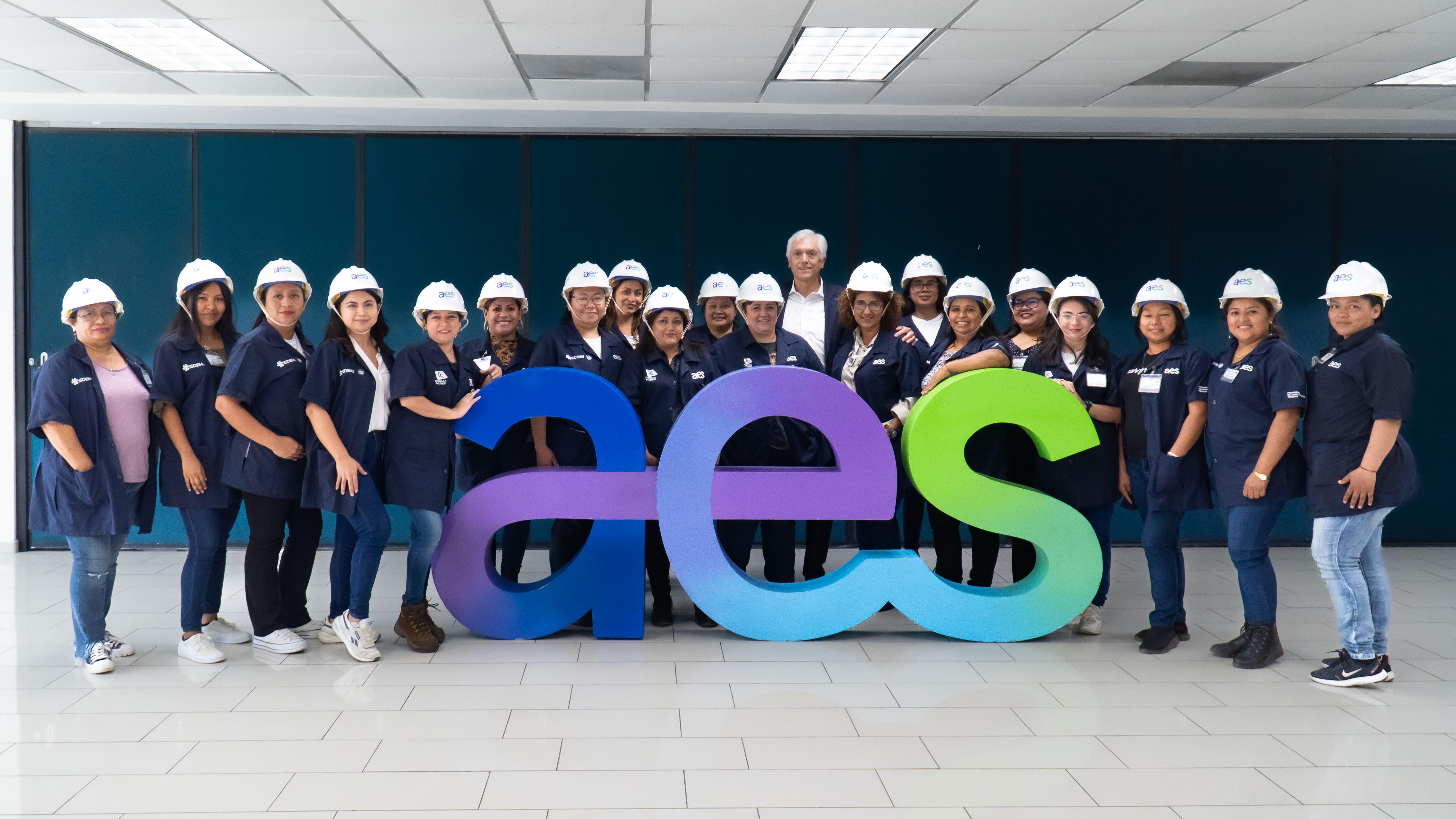 A group of women wearing AES-branded white hard hats and navy-blue uniforms pose for a photo in front of a large, colorful AES logo inside a modern indoor space. They are standing together, smiling confidently, with a man in business attire among them. The group appears to be part of a training or professional development event, representing diversity and inclusion in the electrical industry.