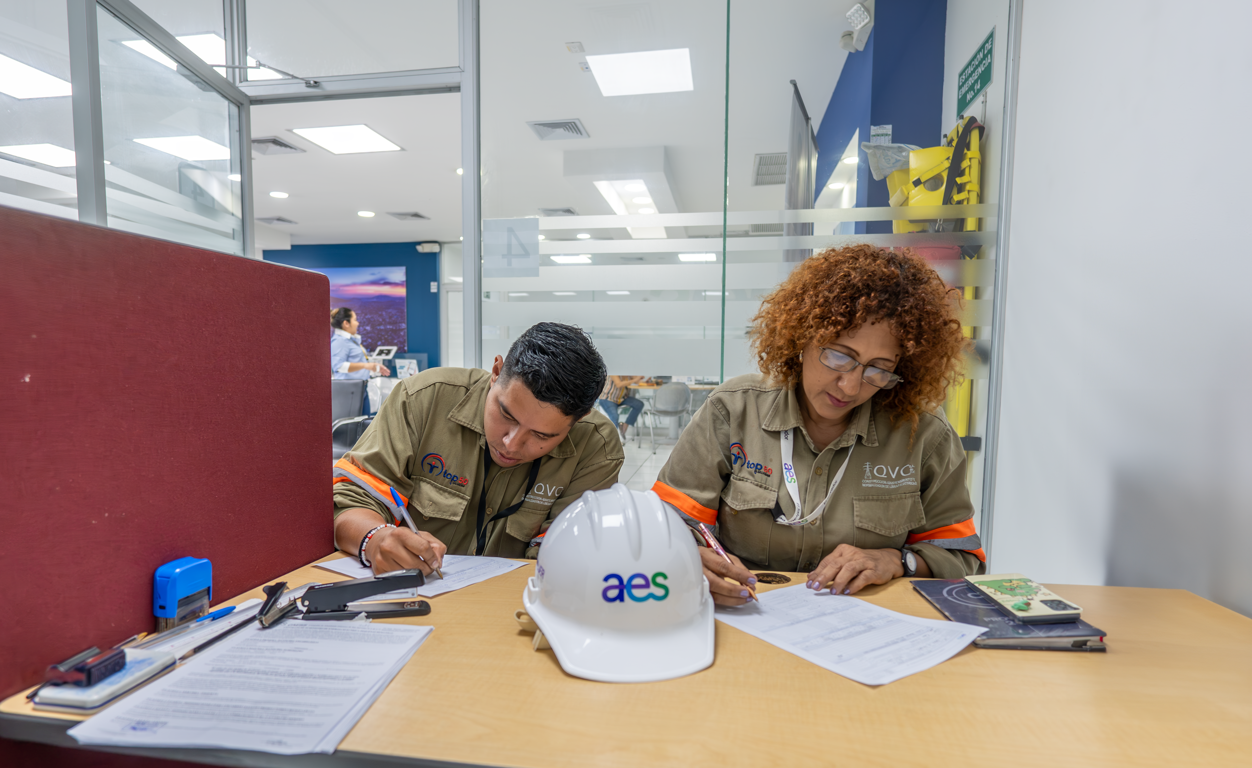 Dos trabajadores con uniformes caqui y gafas están sentados en un escritorio de color claro en una oficina luminosa. Están revisando documentos y escribiendo.