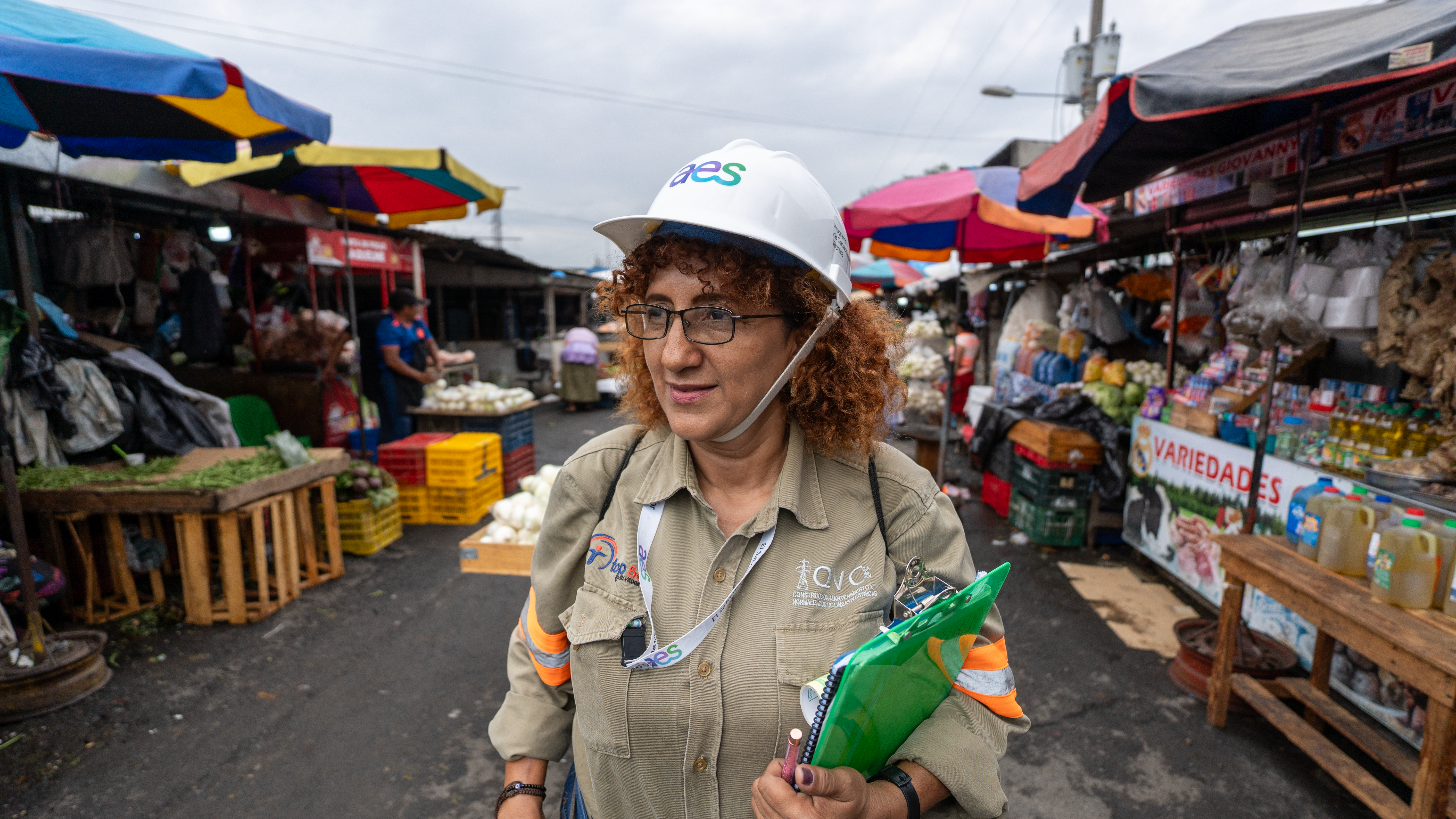 Una mujer adulta con cabello rizado castaño rojizo, gafas y un casco blanco con el logo "aes" está de pie en un mercado al aire libre. Viste una camisa de trabajo caqui con rayas reflectantes naranjas y sostiene un portapapeles verde brillante.