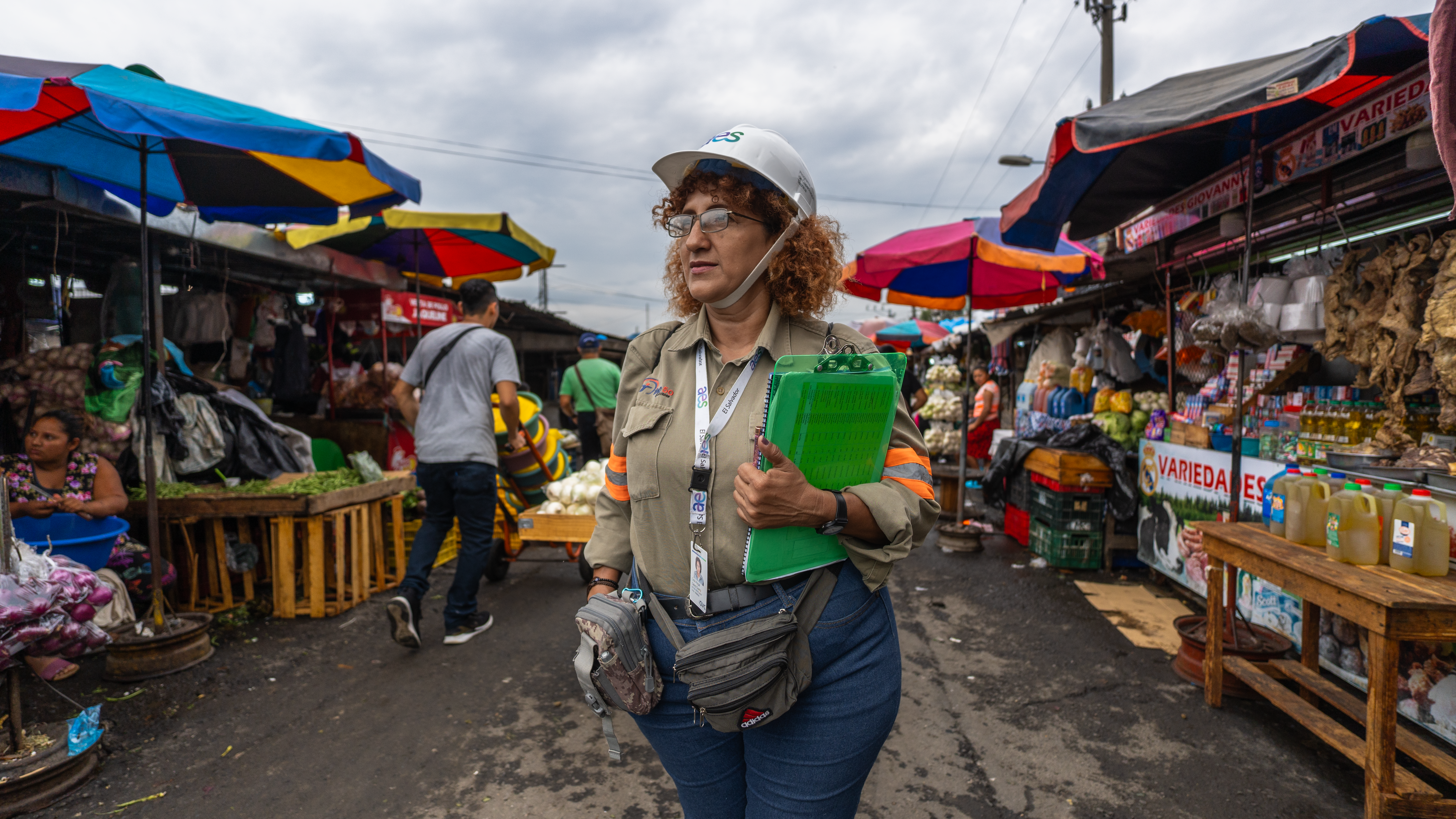 Una mujer con gafas, casco blanco y ropa de trabajo caqui sostiene un portapapeles verde brillante en un mercado al aire libre lleno de gente y puestos coloridos bajo toldos.