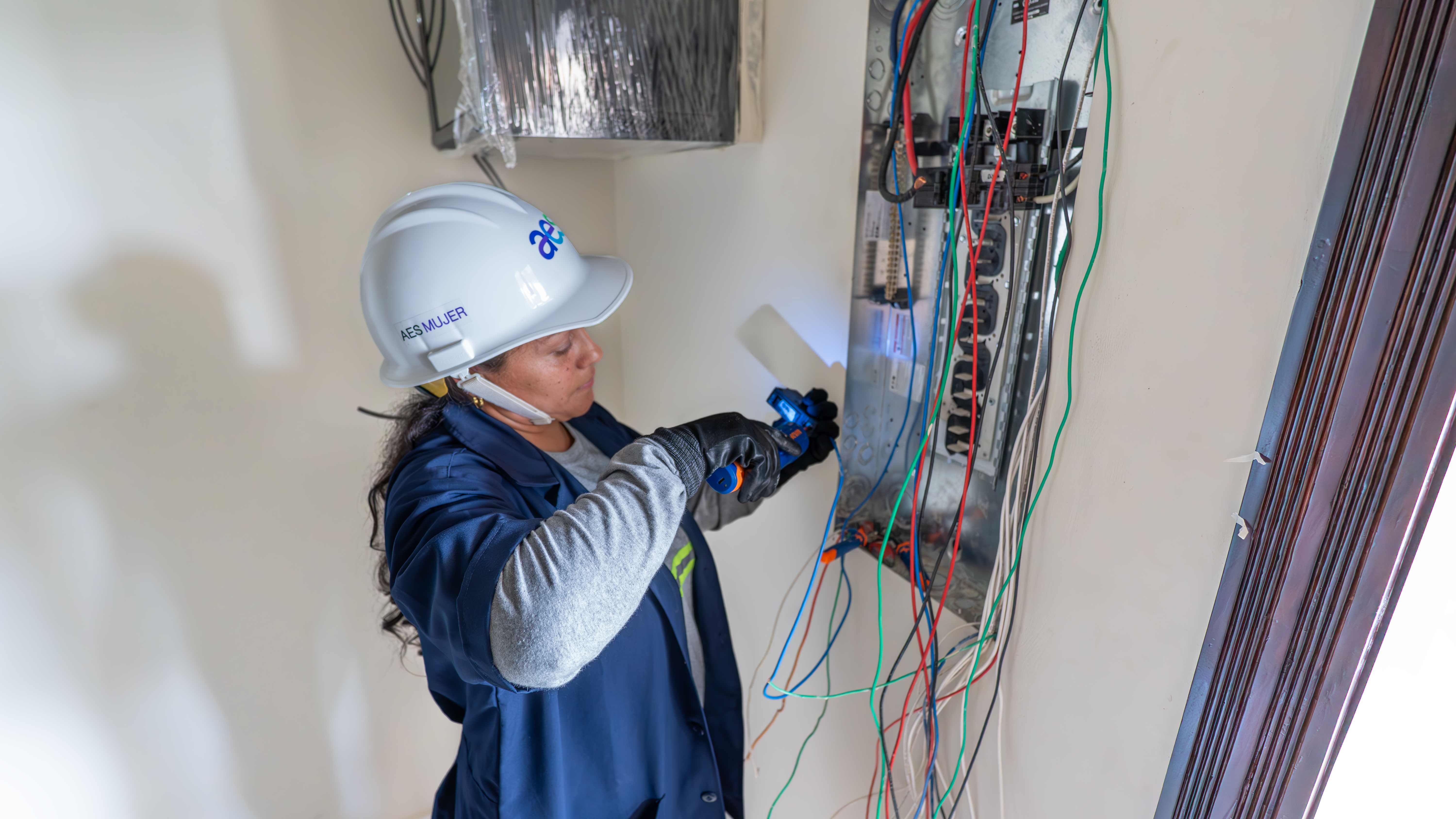 Una mujer electricista con un casco de seguridad blanco y una chaqueta de trabajo azul examina un panel de cableado eléctrico. Sostiene una pequeña herramienta o linterna que emite una luz azul para inspeccionar los cables. Numerosos cables de colores (rojos, verdes, azules, blancos, negros) salen del panel y se extienden hacia la parte inferior del encuadre. Sus manos están protegidas con guantes negros y grises mientras trabaja.