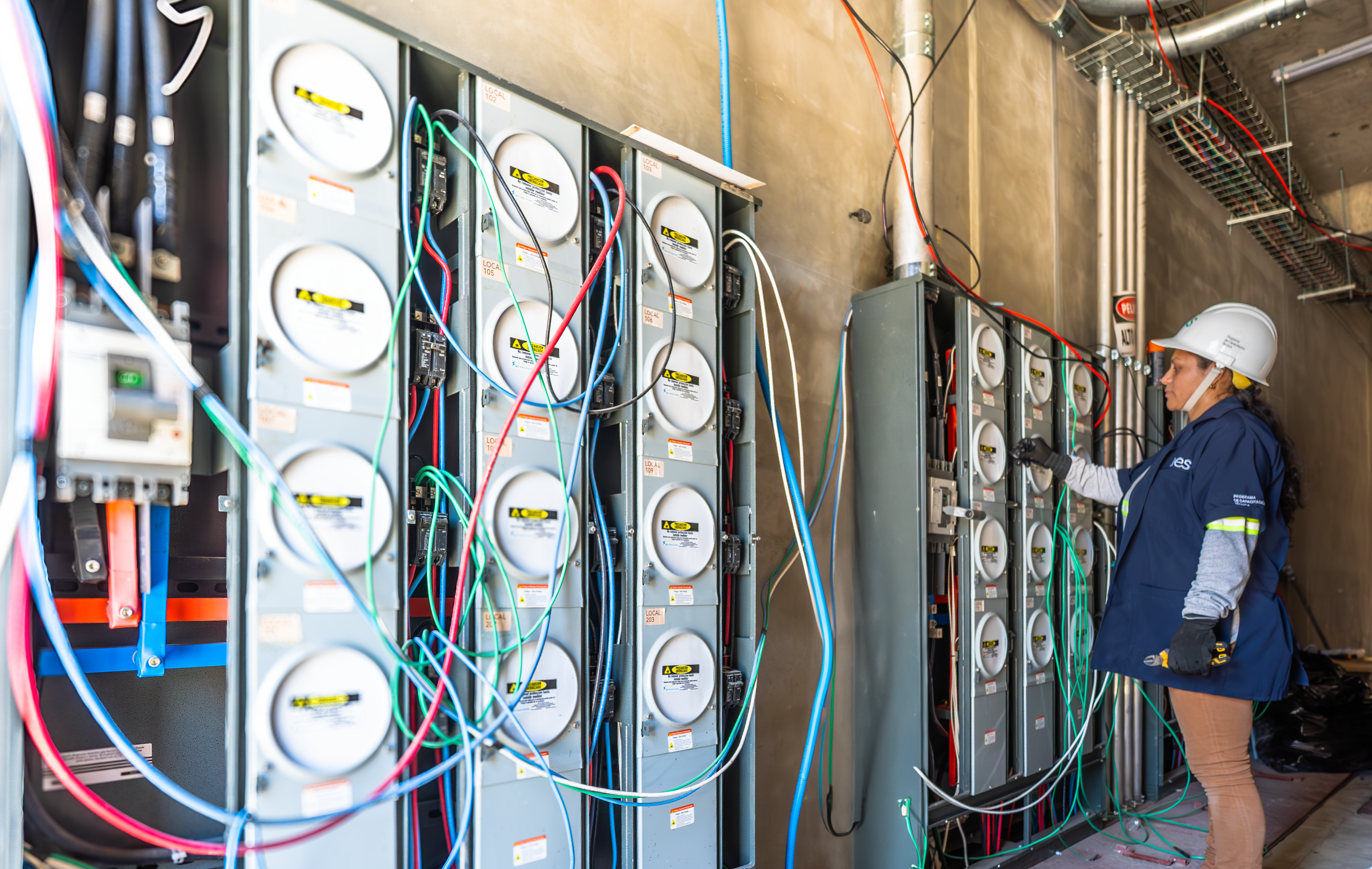 electricista-trabajando-en-medidores-electricos Una electricista con un casco de seguridad blanco y una chaqueta de trabajo azul revisa una hilera de medidores eléctricos en un entorno industrial. Está de pie junto a varios paneles grises interconectados, cada uno con medidores redondos y numerosos cables de colores (rojos, verdes, azules, negros) que sobresalen y se extienden. Parece estar inspeccionando o realizando ajustes en uno de los paneles.