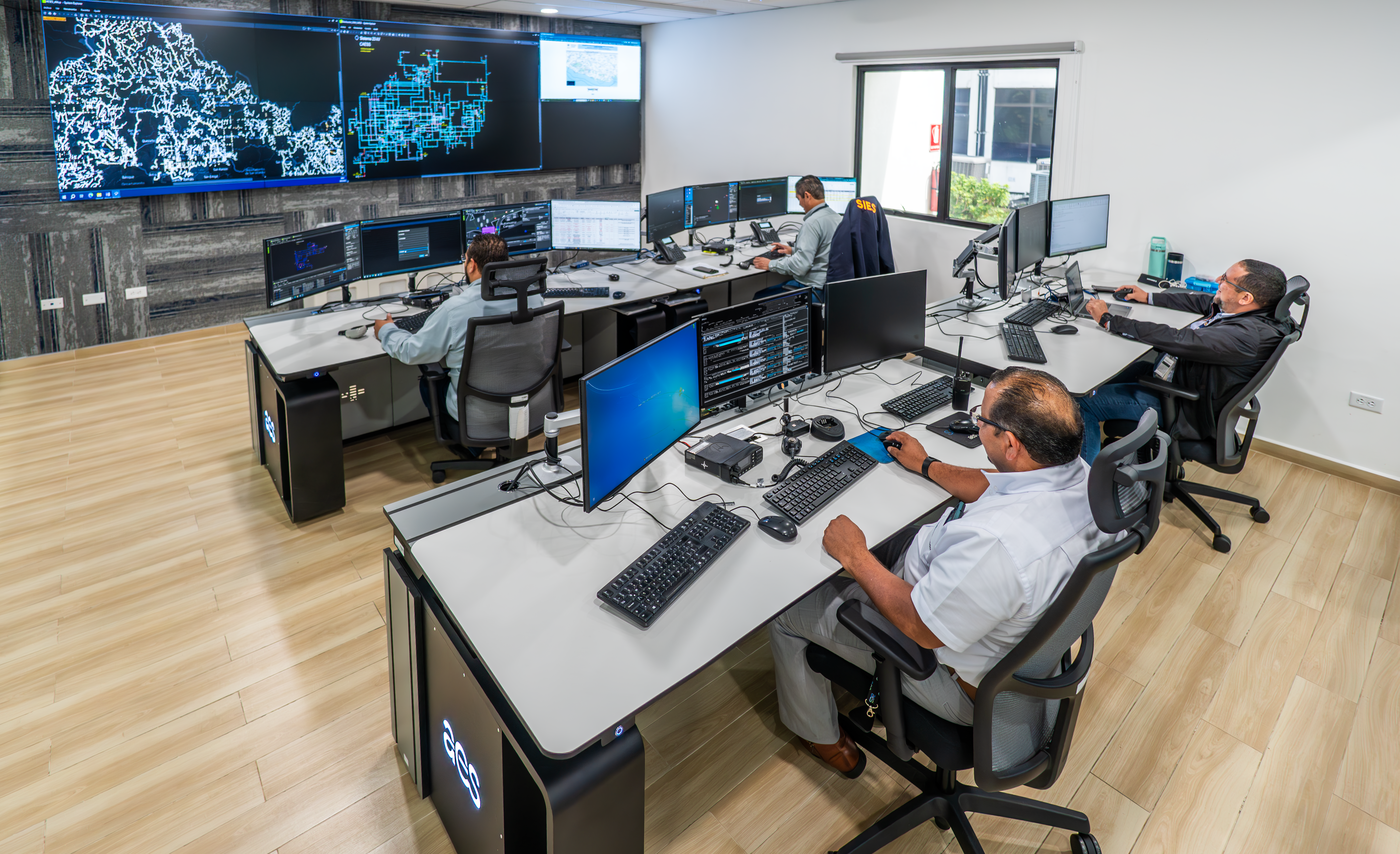 Cuatro ingenieros están sentados en estaciones de trabajo individuales en un centro de control moderno. Cada estación tiene múltiples monitores. En la pared del fondo, se ven dos grandes pantallas que muestran mapas complejos y datos de red. La sala tiene paredes blancas y un piso de madera clara. Los ingenieros están concentrados en sus tareas, algunos usando teclados y ratones. Hay escritorios negros y bases de computadora con un logo similar a "aes". Fuentes