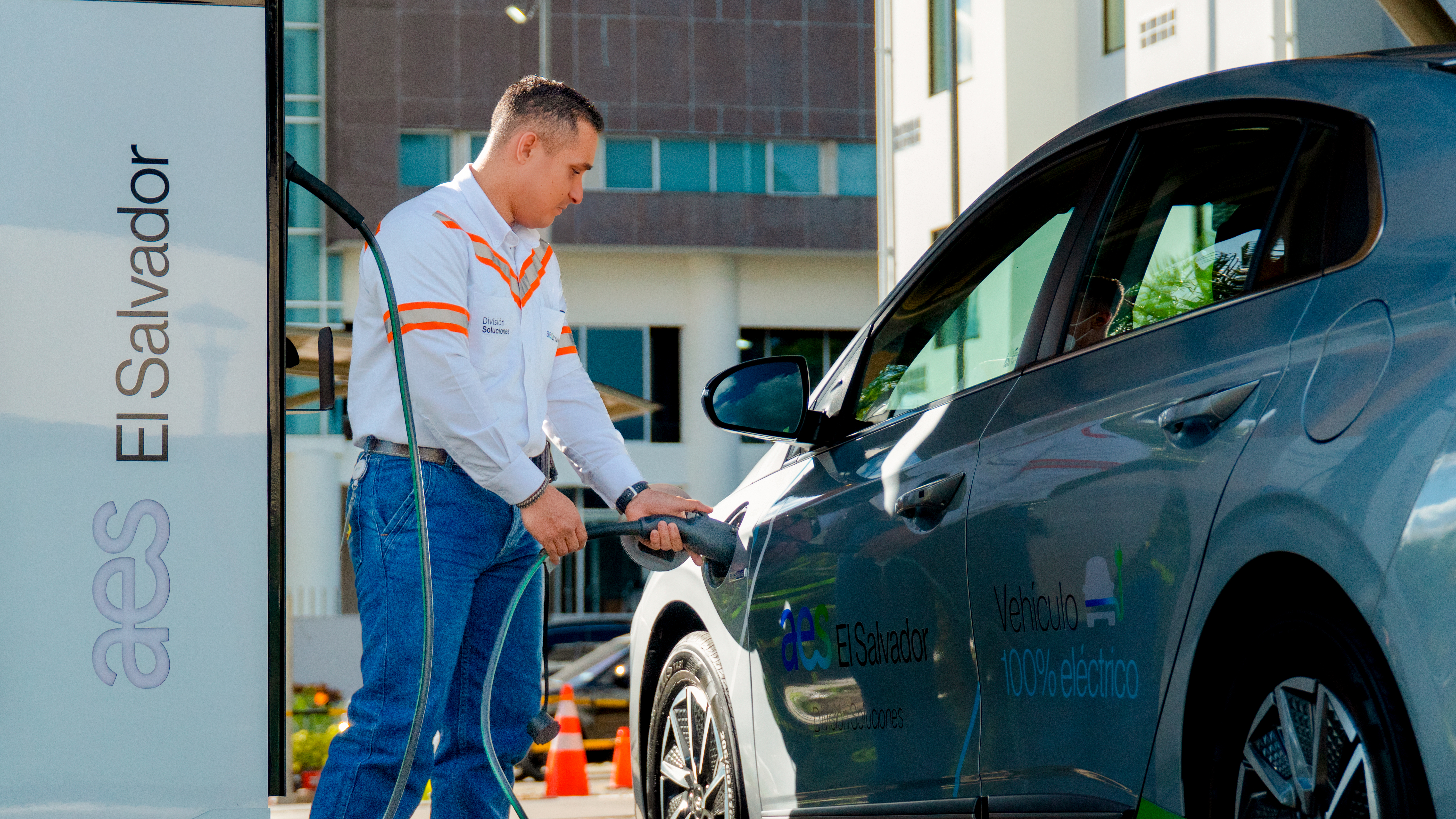 Un hombre conecta una manguera de carga a un coche eléctrico gris. El coche tiene la etiqueta "Vehículo 100% Eléctrico" y el logo de "El Salvador". El hombre viste una camisa blanca con detalles naranjas y pantalones vaqueros. Está de pie junto a una estación de carga blanca con el logo "aes El Salvador"