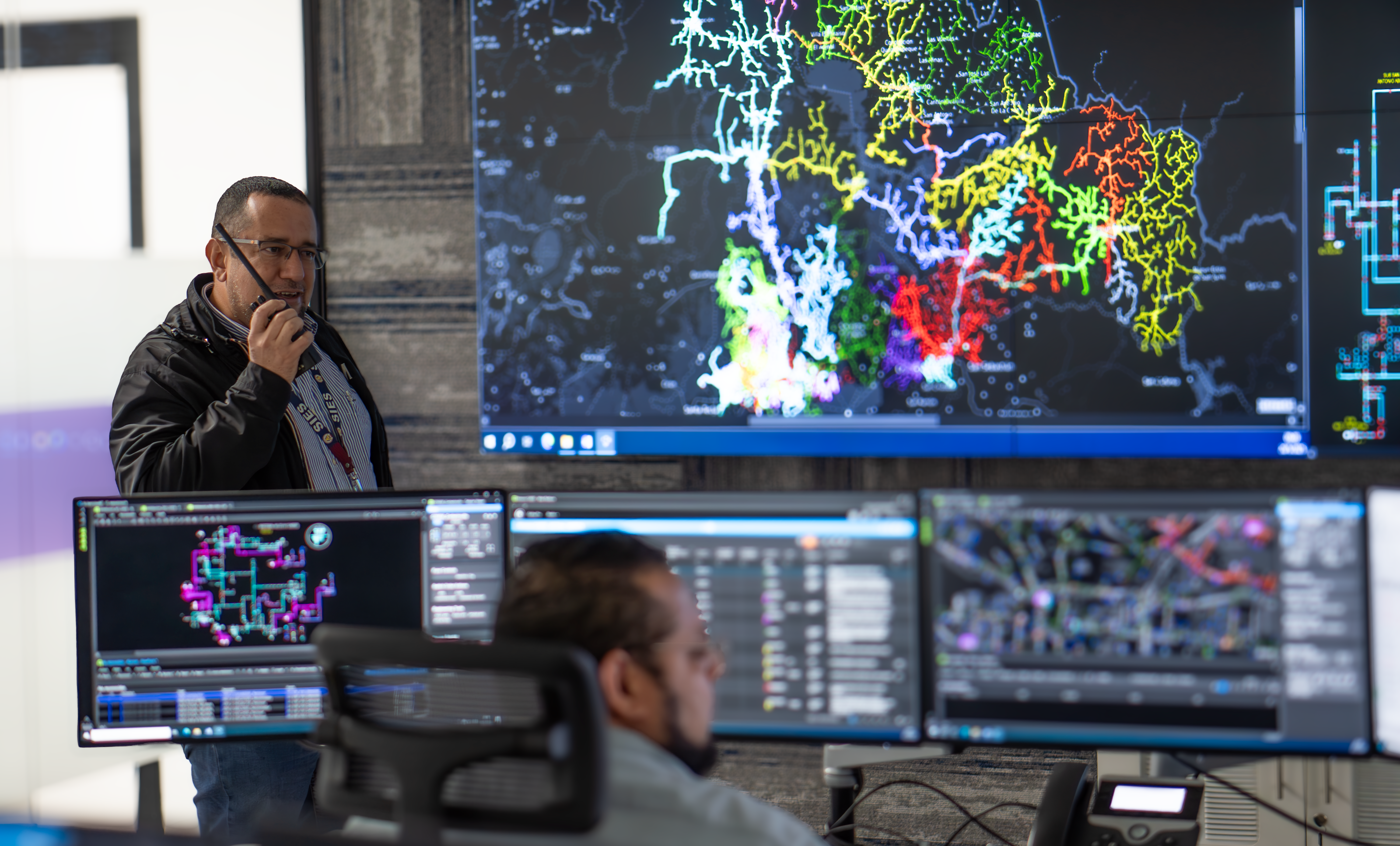 Un hombre con gafas y una chaqueta negra está de pie en un centro de control, hablando por un radio. Detrás de él, una gran pantalla muestra un mapa detallado con líneas de colores, que probablemente representa una red eléctrica. Delante de él, en primer plano y desenfocado, se ve la parte trasera de la cabeza de otro hombre sentado frente a múltiples monitores que muestran más gráficos y datos de la red. La escena sugiere un entorno de monitoreo y gestión de operaciones.