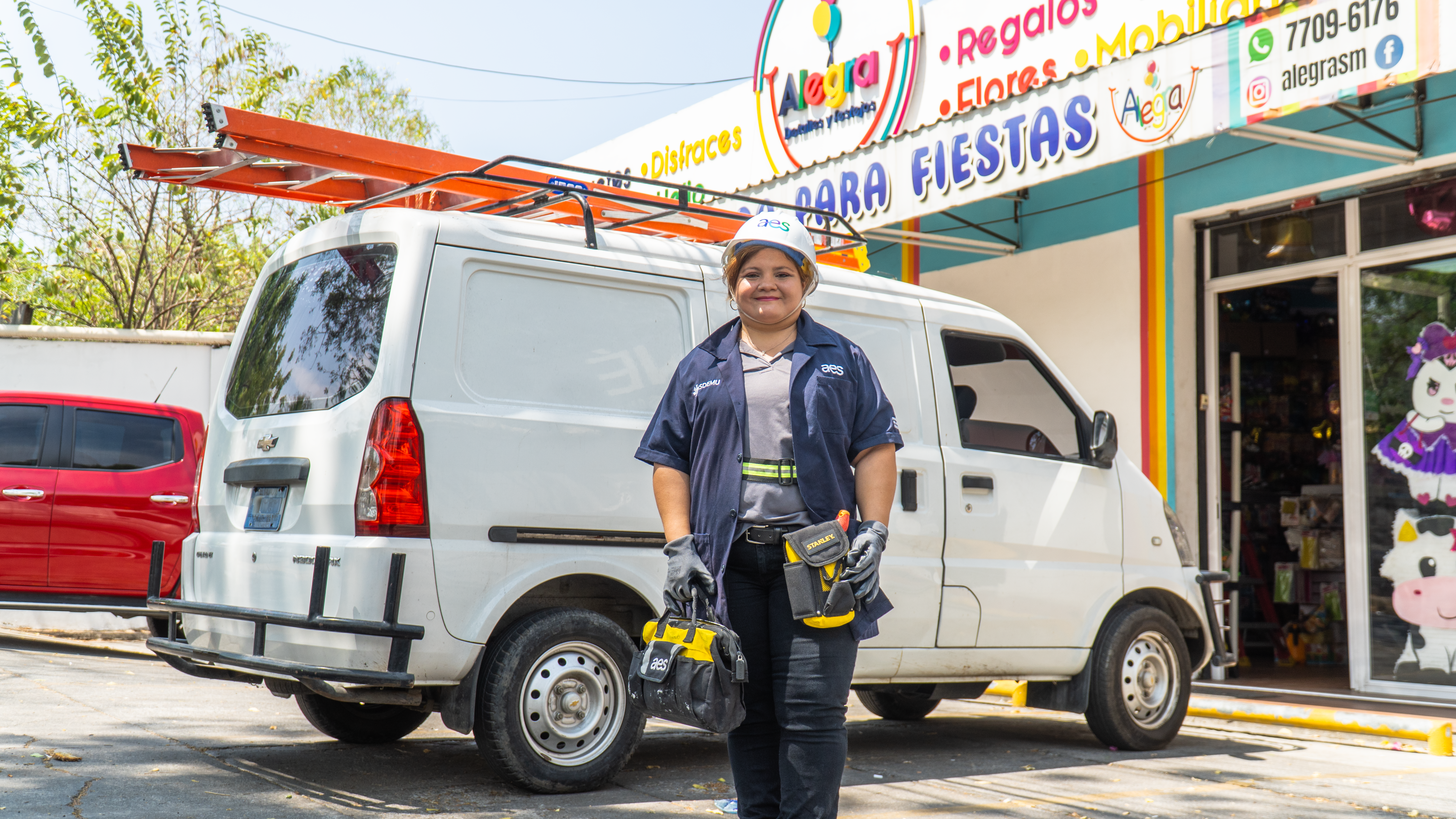 Una mujer sonriente con casco blanco, camisa azul oscuro, pantalón oscuro y guantes grises está de pie frente a una camioneta blanca con una escalera naranja en el techo. Ella sostiene una bolsa de herramientas amarilla y negra en su mano izquierda y una herramienta amarilla en la derecha.