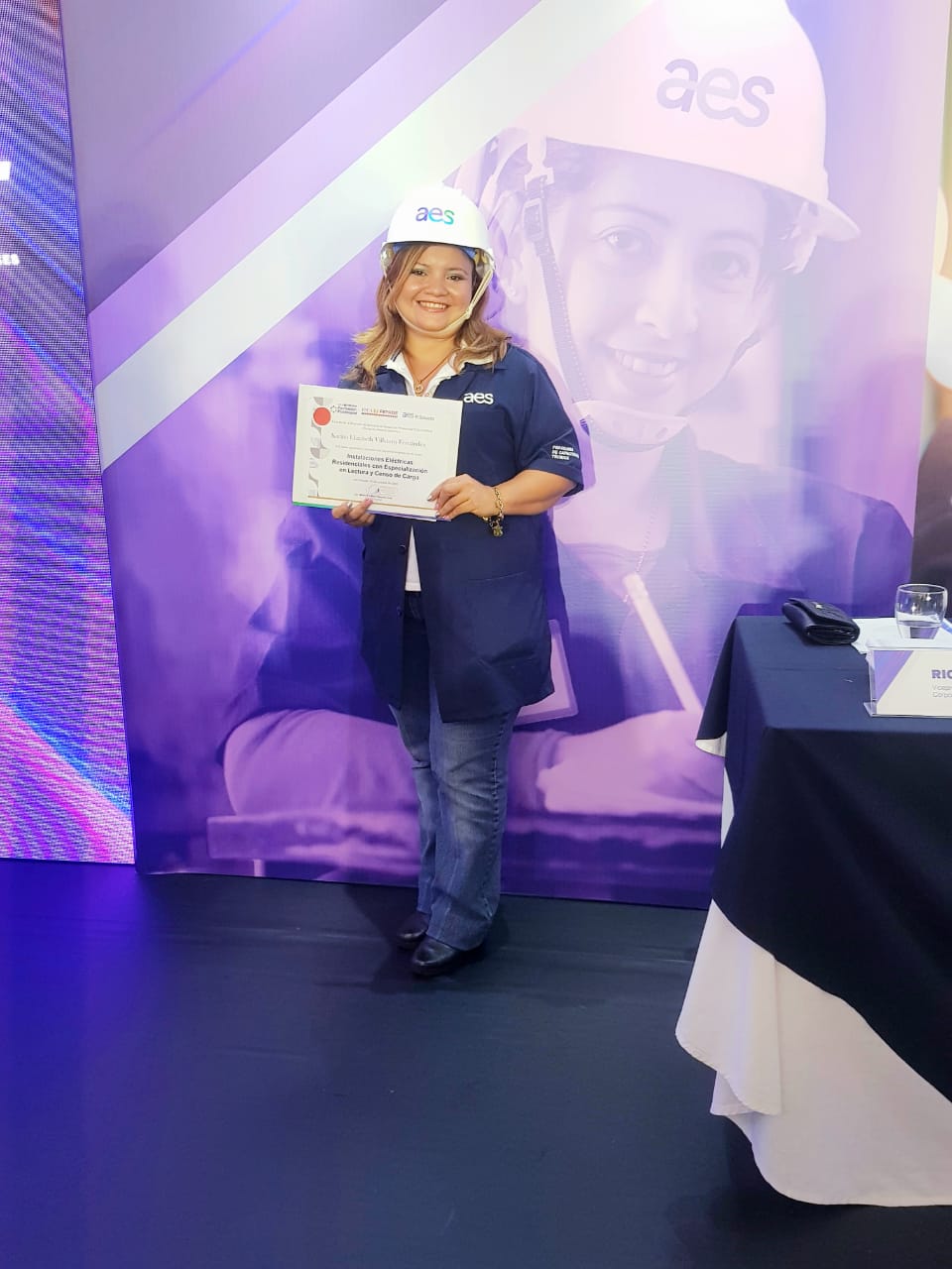 mujer-casco-certificado-evento Una mujer sonriente con casco blanco con el logo "aes", camisa azul oscuro y jeans está de pie y sostiene un certificado en sus manos. Ella está frente a un fondo morado con la imagen borrosa de otra mujer con casco.