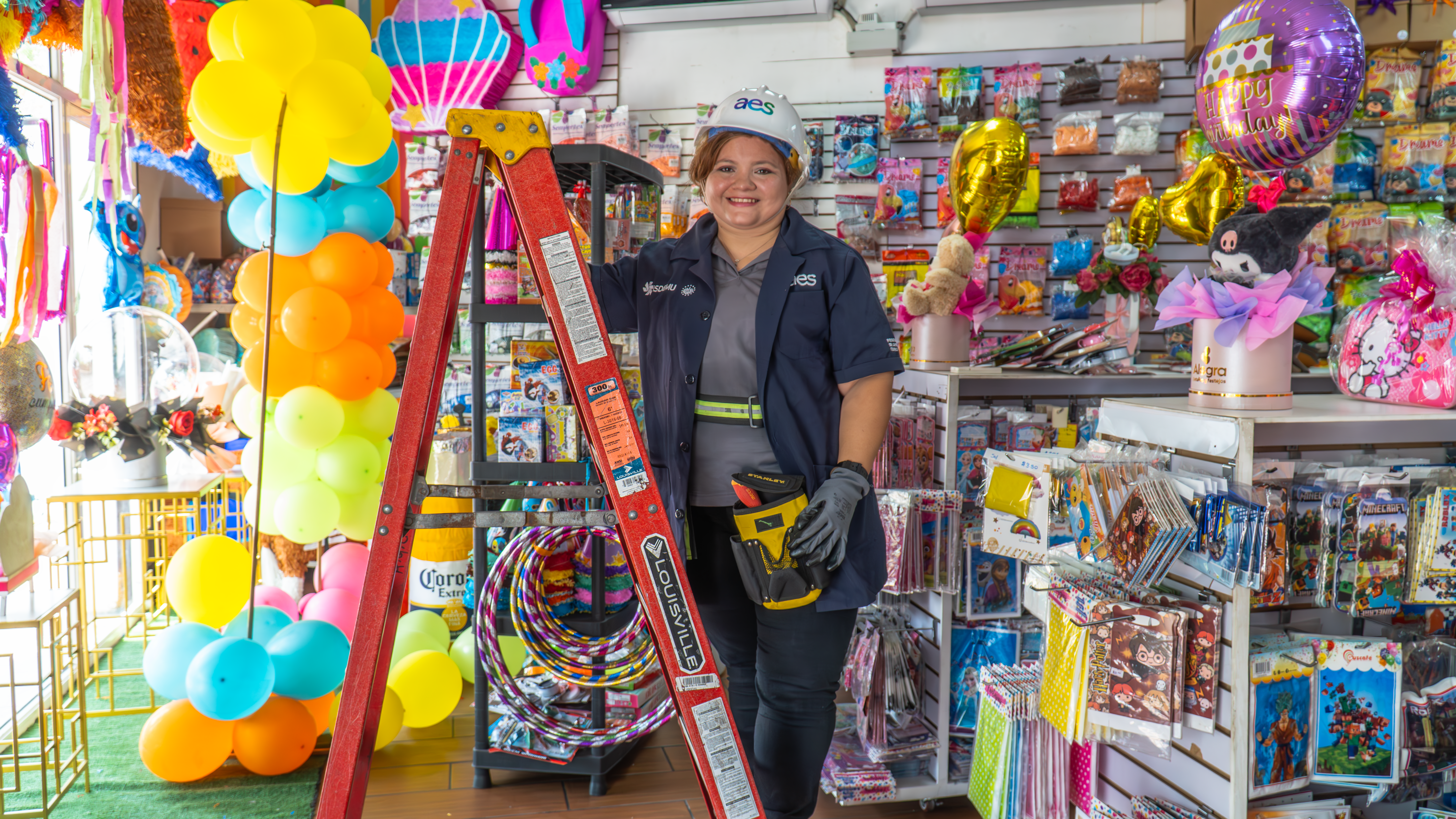 Una mujer sonriente con casco blanco, camisa azul oscuro y guantes grises está de pie junto a una escalera roja en una tienda de artículos para fiestas llena de decoraciones coloridas como globos y piñatas. Ella sostiene una herramienta amarilla y negra en su mano derecha.