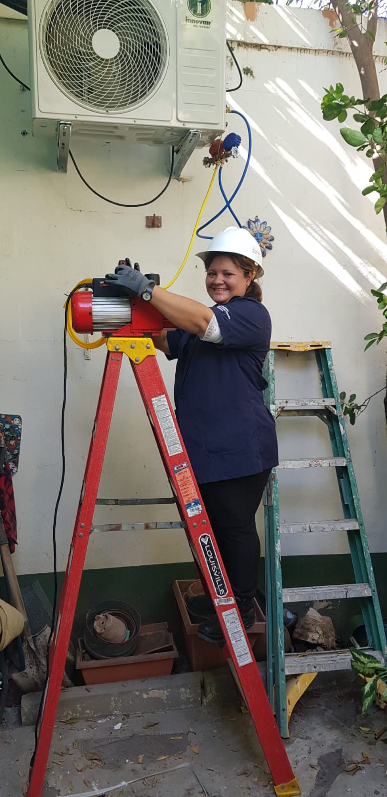 Una mujer con casco blanco, camisa azul oscuro y guantes grises está de pie en una escalera roja trabajando en una unidad de aire acondicionado blanca montada en una pared exterior blanca. Hay otra escalera verde apoyada contra la pared a su derecha y vegetación visible en los bordes.