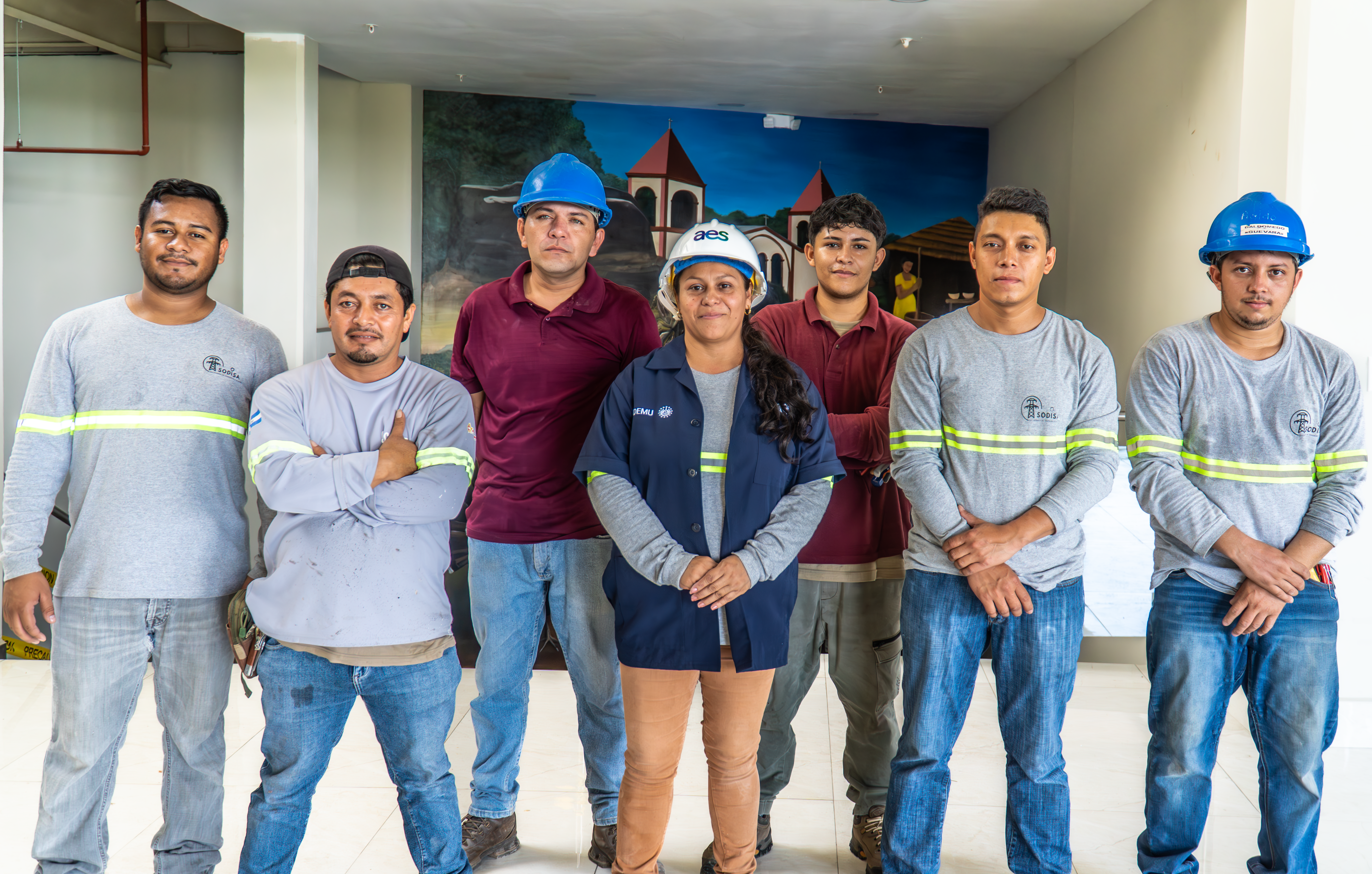 Un grupo diverso de siete trabajadores de la construcción (seis hombres y una mujer) posan de pie en el interior frente a un mural que representa edificios y una persona. La mujer en el centro usa un casco blanco y una chaqueta azul con el logotipo "aes".