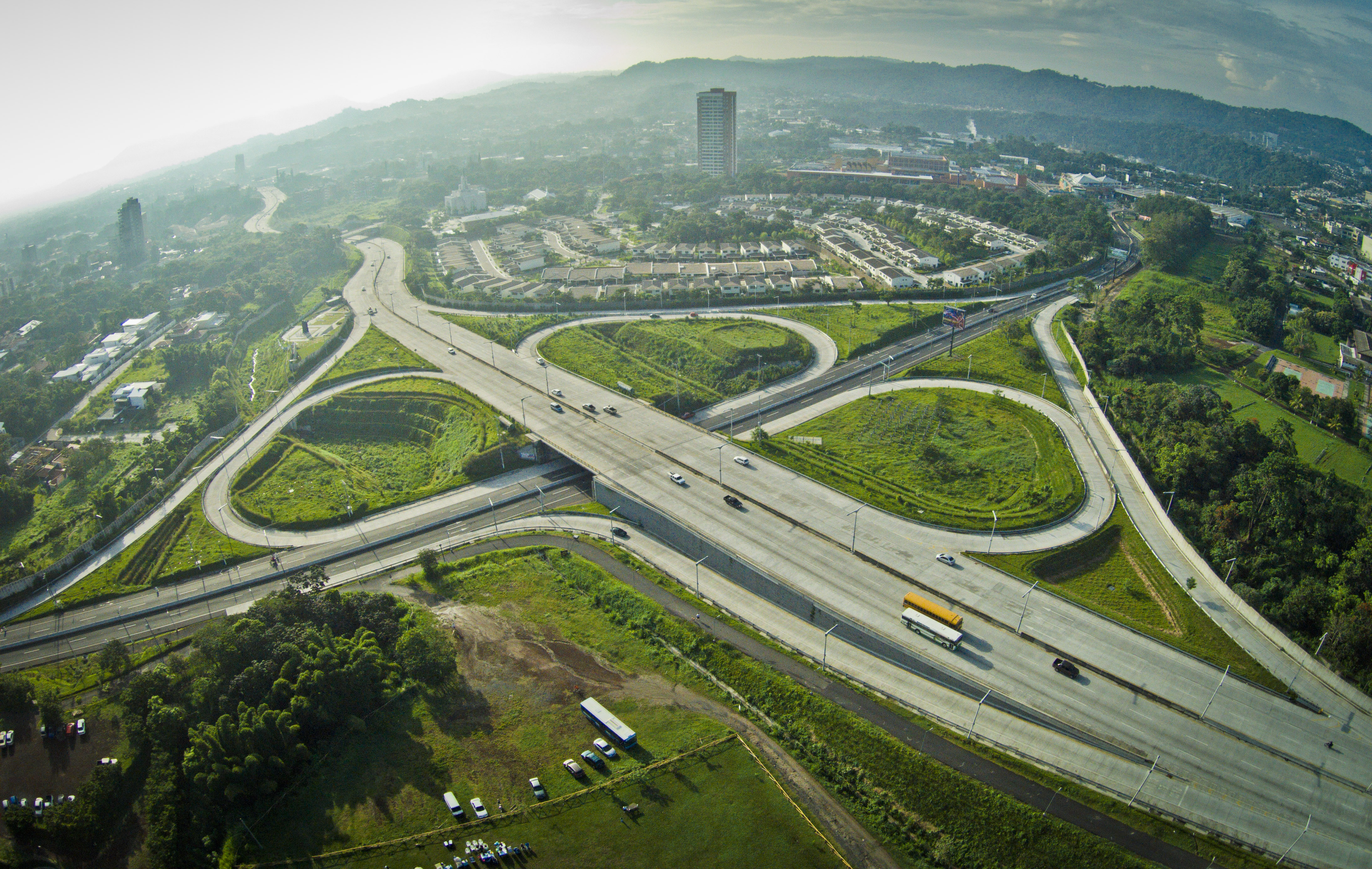 Vista aérea de un gran intercambio de autopistas con múltiples rampas curvas que se elevan sobre el paisaje circundante. Se ven varios vehículos viajando por las carreteras. Al fondo, se extiende una ciudad con edificios y zonas residenciales, con montañas verdes y onduladas que se elevan en la distancia bajo un cielo nublado.