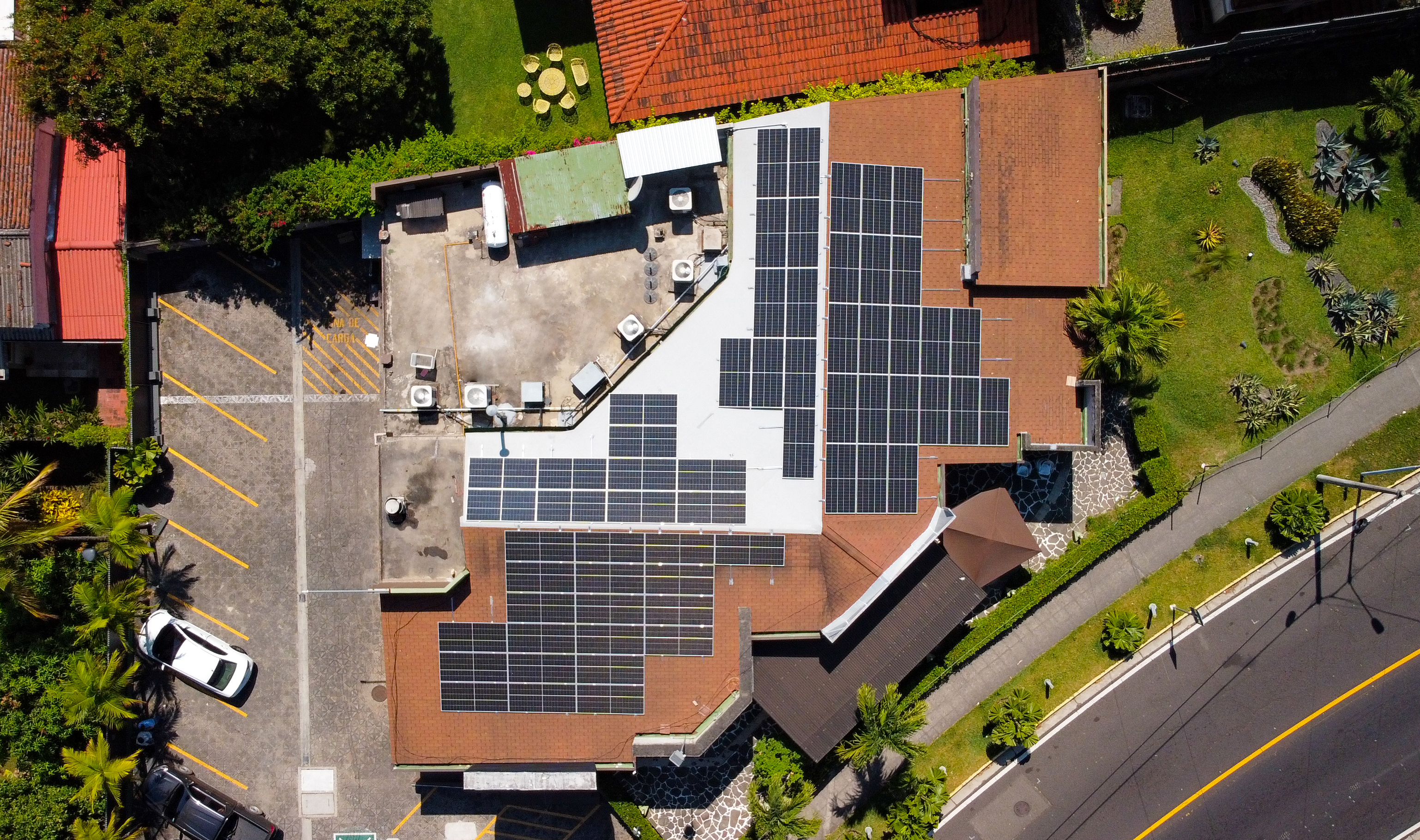 Vista aérea de un tejado cubierto con numerosos paneles solares dispuestos en varias secciones. El edificio de abajo tiene un aparcamiento adyacente con algunos coches estacionados y está rodeado de vegetación, incluyendo árboles y jardines. Una calle con un coche en movimiento bordea la propiedad.