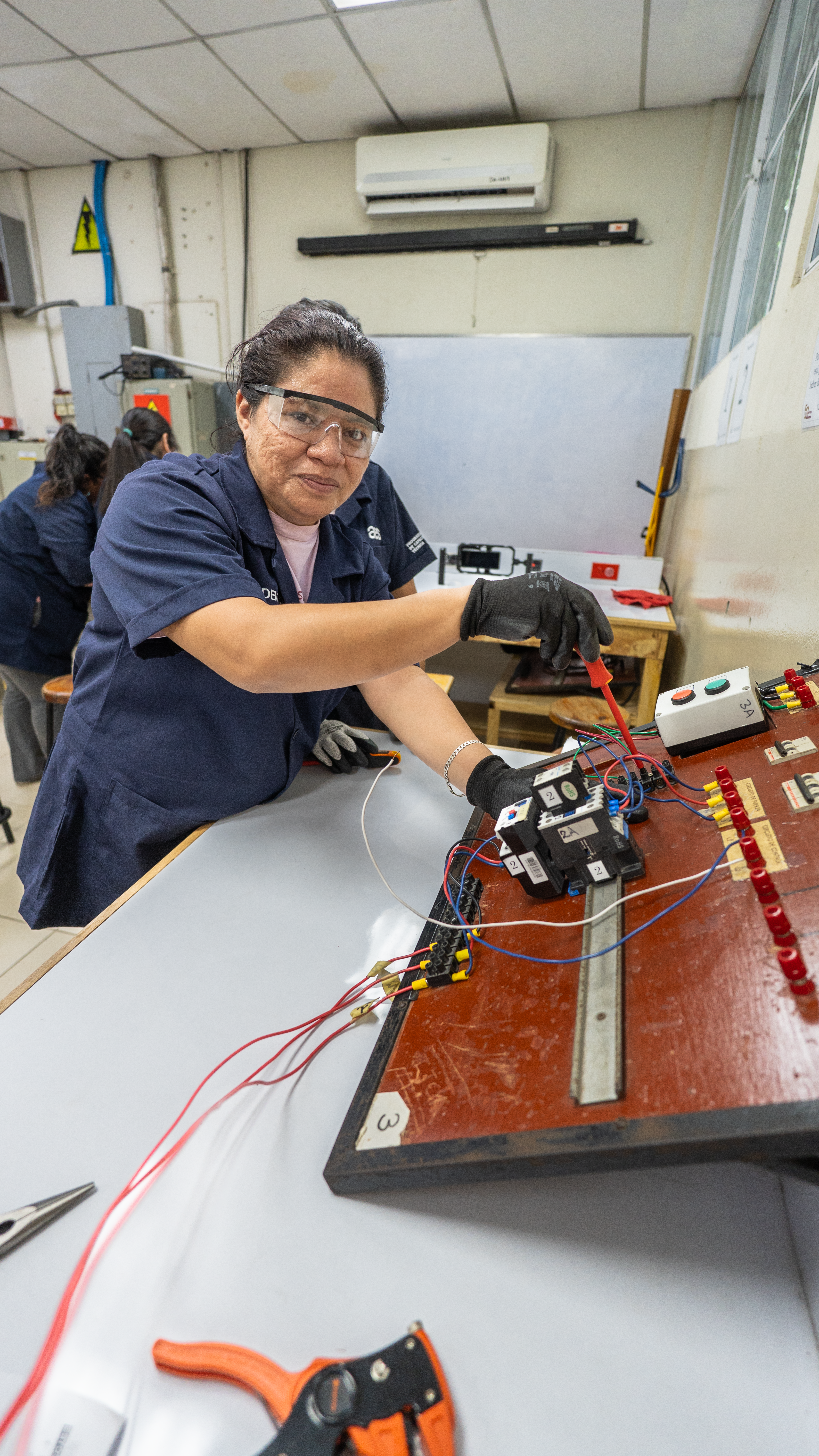 Una mujer con gafas de seguridad y guantes negros conecta cables de colores a un panel electrónico marrón con componentes y luces. Está inclinada sobre una mesa gris, utilizando una herramienta roja y negra. En el fondo, desenfocado, se ven otras personas trabajando en mesas similares en un aula con paredes blancas, un pizarrón blanco y un aire acondicionado en la parte superior.