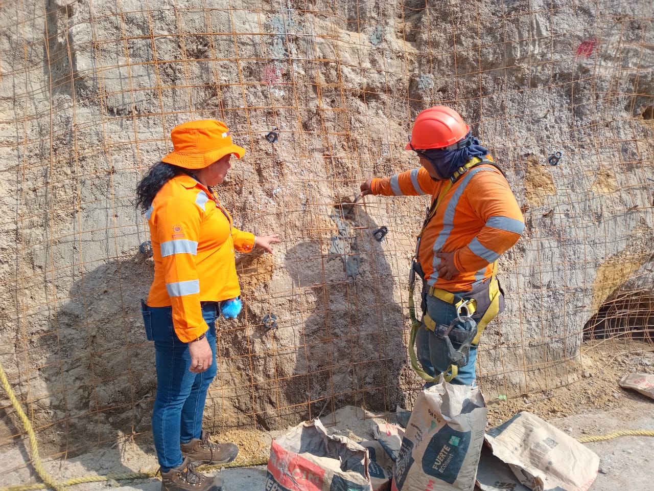 Dos trabajadores de la construcción con ropa de seguridad naranja y cascos inspeccionan una pared de roca. La persona de la izquierda, que lleva un sombrero naranja en lugar de casco, señala la pared, mientras que la de la derecha, con casco, examina la superficie rocosa que parece estar reforzada con malla metálica.