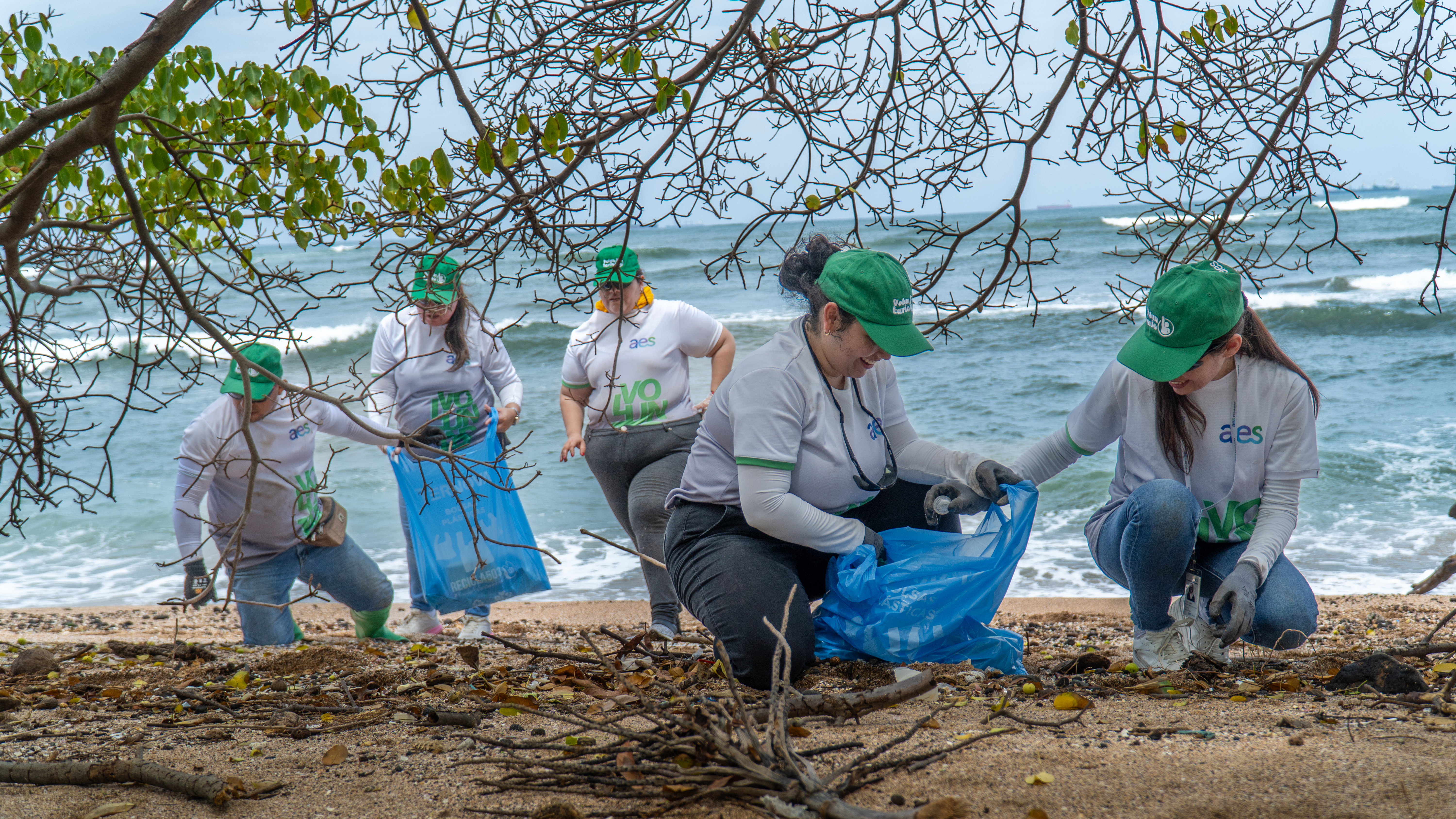 Un grupo de cinco personas, la mayoría con camisetas blancas, gorras verdes y guantes, están en una playa, recogiendo basura en bolsas de plástico azules. Se agachan y manipulan objetos del suelo. Al fondo, el océano tiene olas suaves y el cielo es visible, aunque ligeramente nublado.