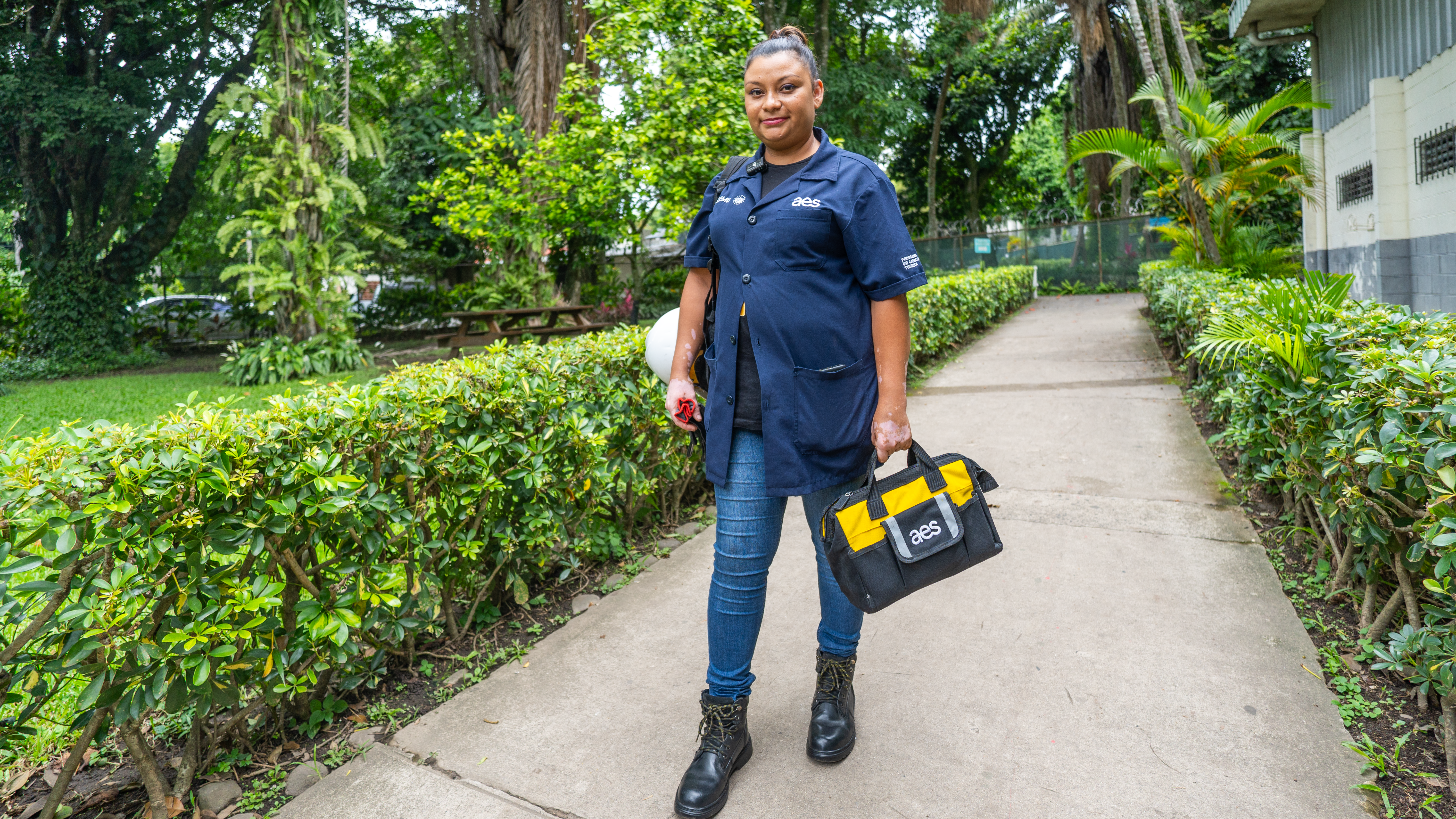 Una mujer joven, vestida con una camisa de trabajo azul oscuro con el logo "aes" en el pecho, jeans y botas de trabajo negras, camina por un camino de concreto rodeado de setos verdes y árboles altos. Lleva un bolso de herramientas negro y amarillo con el logo "aes" en su mano izquierda y un casco de seguridad blanco en su mano derecha.