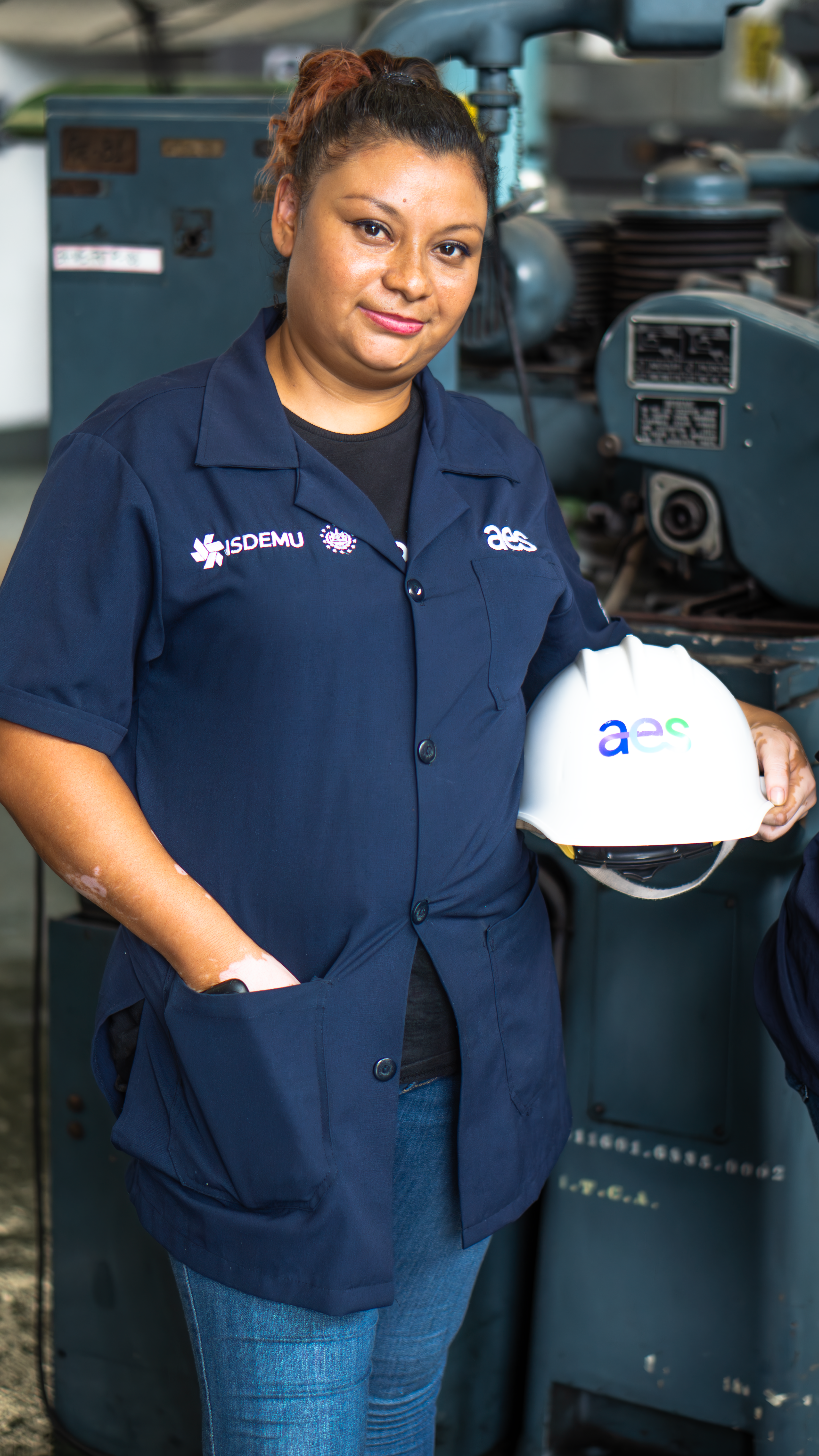 Una mujer joven con una camisa de trabajo azul oscuro con el logo "aes" a la derecha, y jeans, posa sosteniendo un casco de seguridad blanco con el logo "aes" en un entorno industrial.