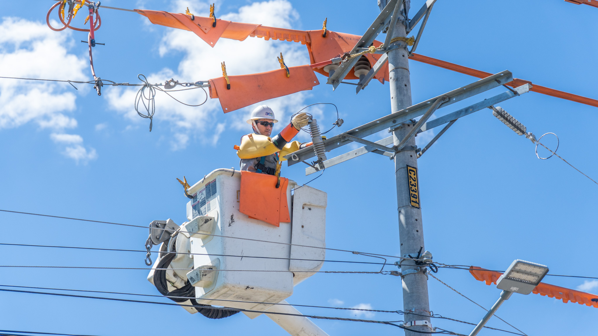 Un trabajador de servicios públicos con casco y equipo de seguridad se encuentra en una cubeta elevadora, trabajando en un poste de electricidad con varias líneas. El cielo está azul y hay algunas nubes blancas. Se han colocado cubiertas de seguridad de color naranja brillante en las líneas eléctricas cercanas. Debajo del poste, hay otras líneas más delgadas y lo que parece ser una farola.