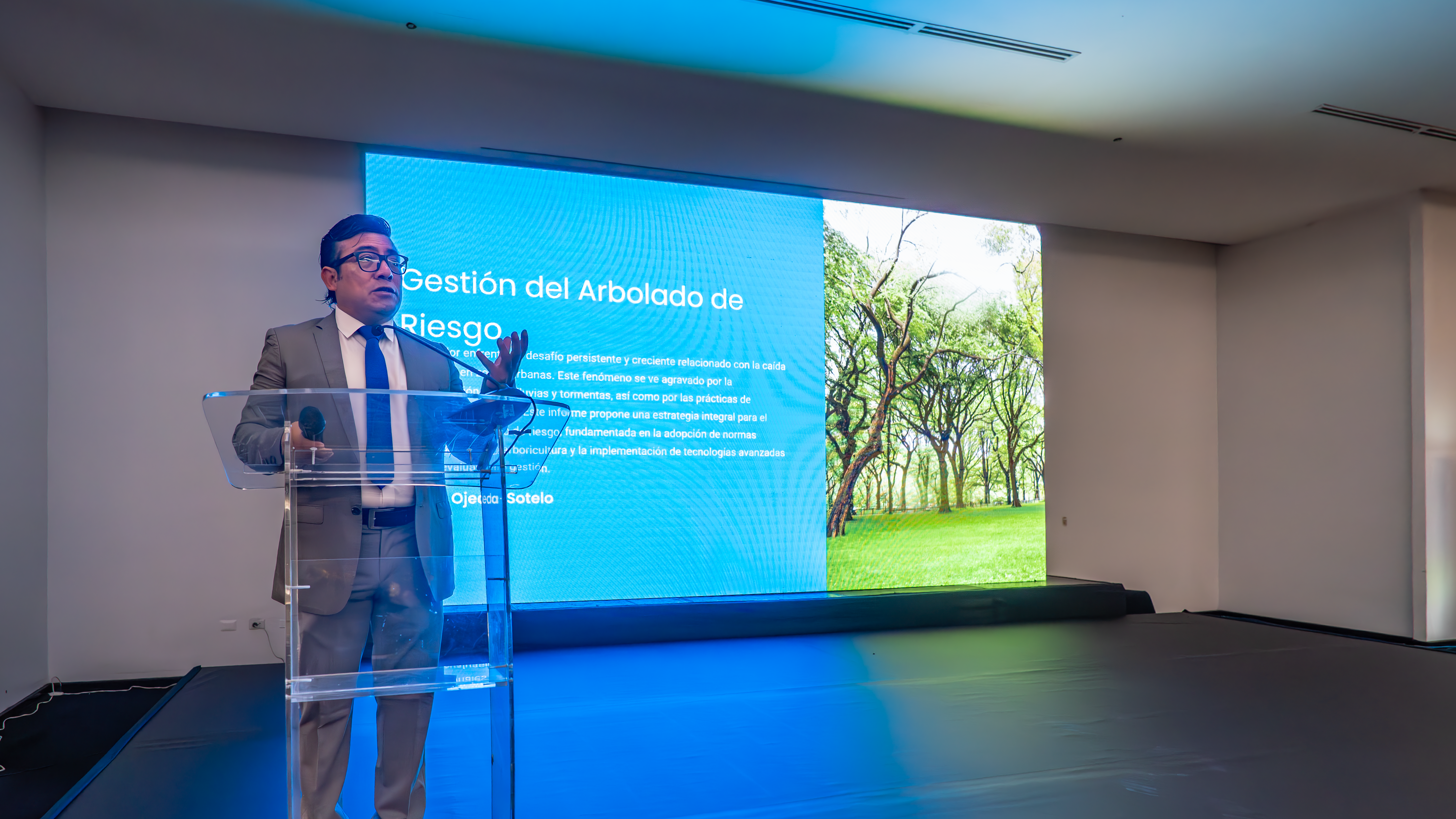 Un hombre con traje gris y gafas está de pie en un podio transparente, gesticulando mientras presenta. Detrás de él hay una gran pantalla que muestra el título "Gestión del Arbolado de Riesgo" en el lado izquierdo y una imagen de árboles con follaje verde en el lado derecho. La pantalla parece ser una pantalla LED o una proyección, y el hombre se encuentra en un escenario oscuro frente a una pared blanca. El lado izquierdo de la imagen está ligeramente más oscuro que el derecho.