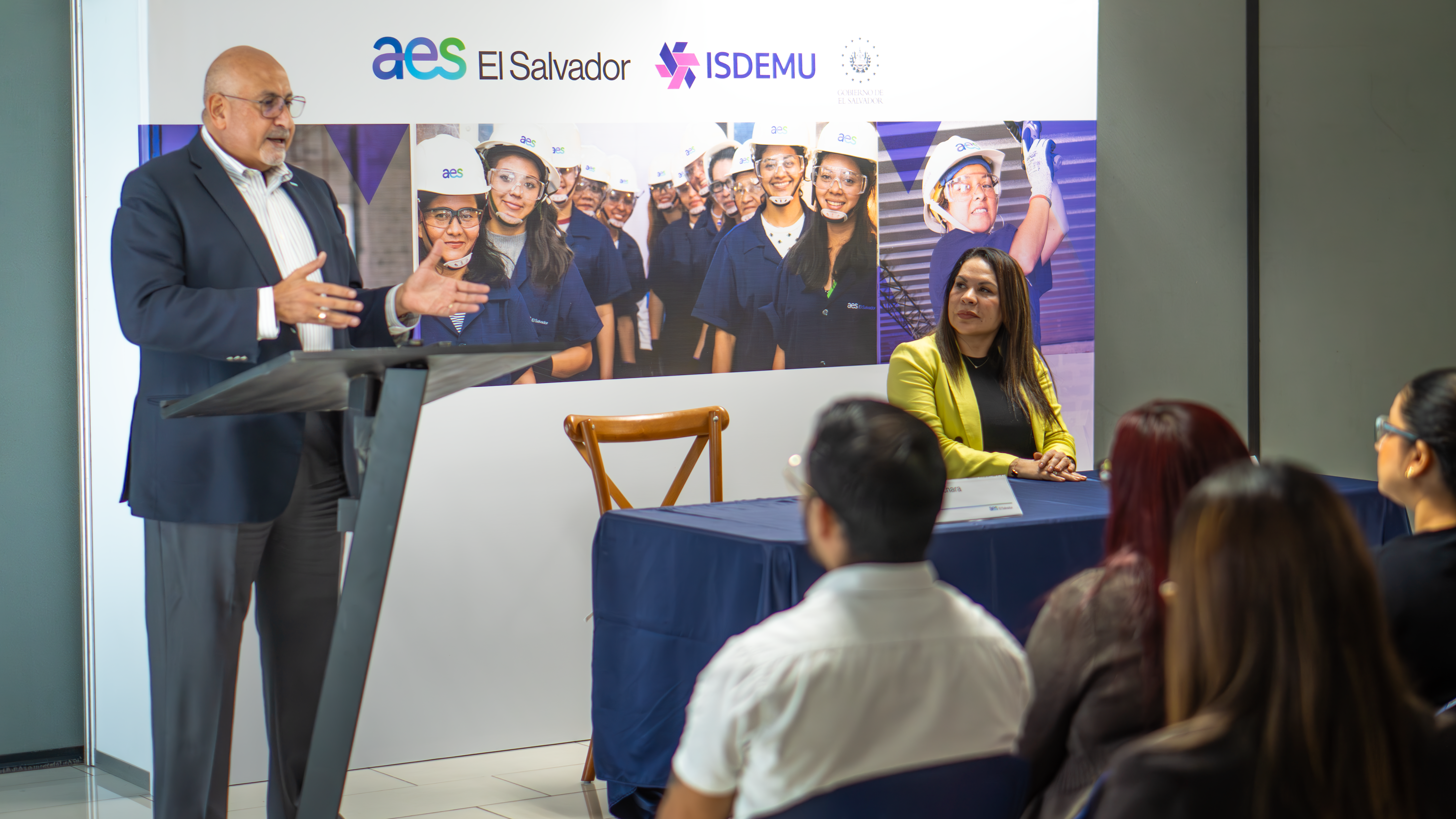 Un hombre calvo con gafas, camisa blanca y traje oscuro está de pie en un podio, gesticulando con ambas manos mientras habla. Detrás de él hay un gran cartel con los logotipos de "aes El Salvador" e "ISDEMU" en la parte superior. El cartel muestra imágenes de varias mujeres jóvenes sonriendo y vistiendo cascos de seguridad y uniformes de trabajo.