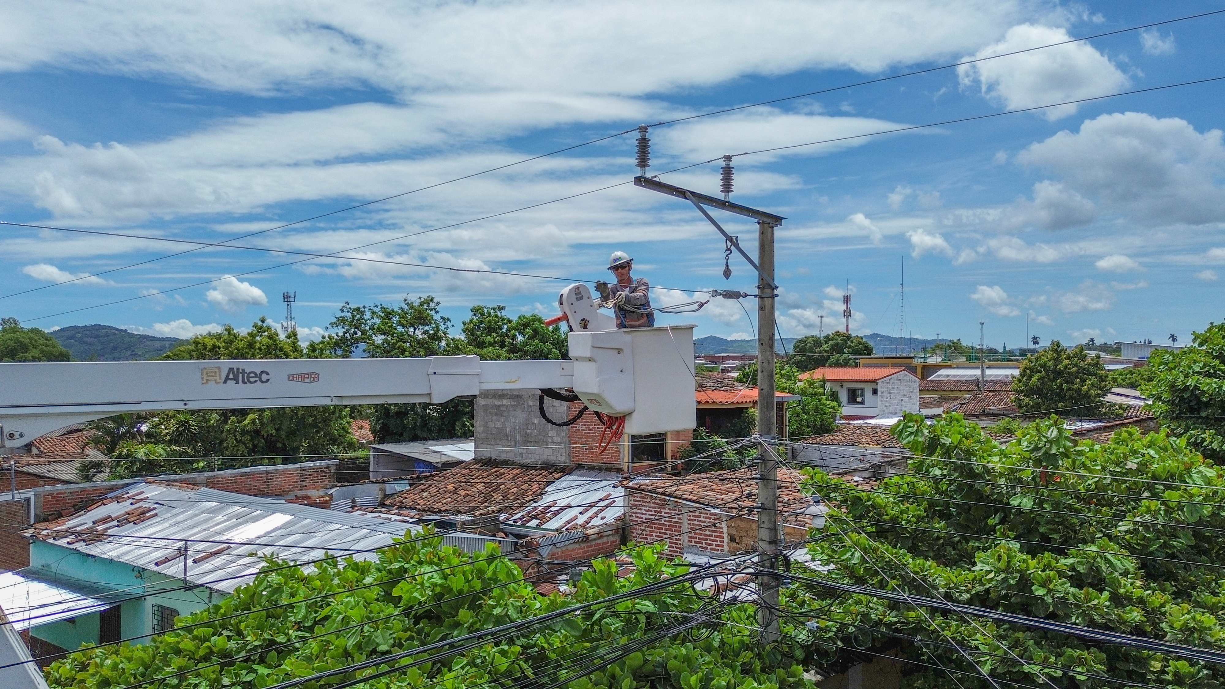 Un trabajador de servicios públicos con casco y equipo de seguridad opera una grúa de la marca Altec en lo alto de un poste de luz, inspeccionando un aislante y cables. La grúa está elevada sobre los tejados de un pueblo con casas de tejas rojas y blancas, y abundantes árboles de hojas verdes.