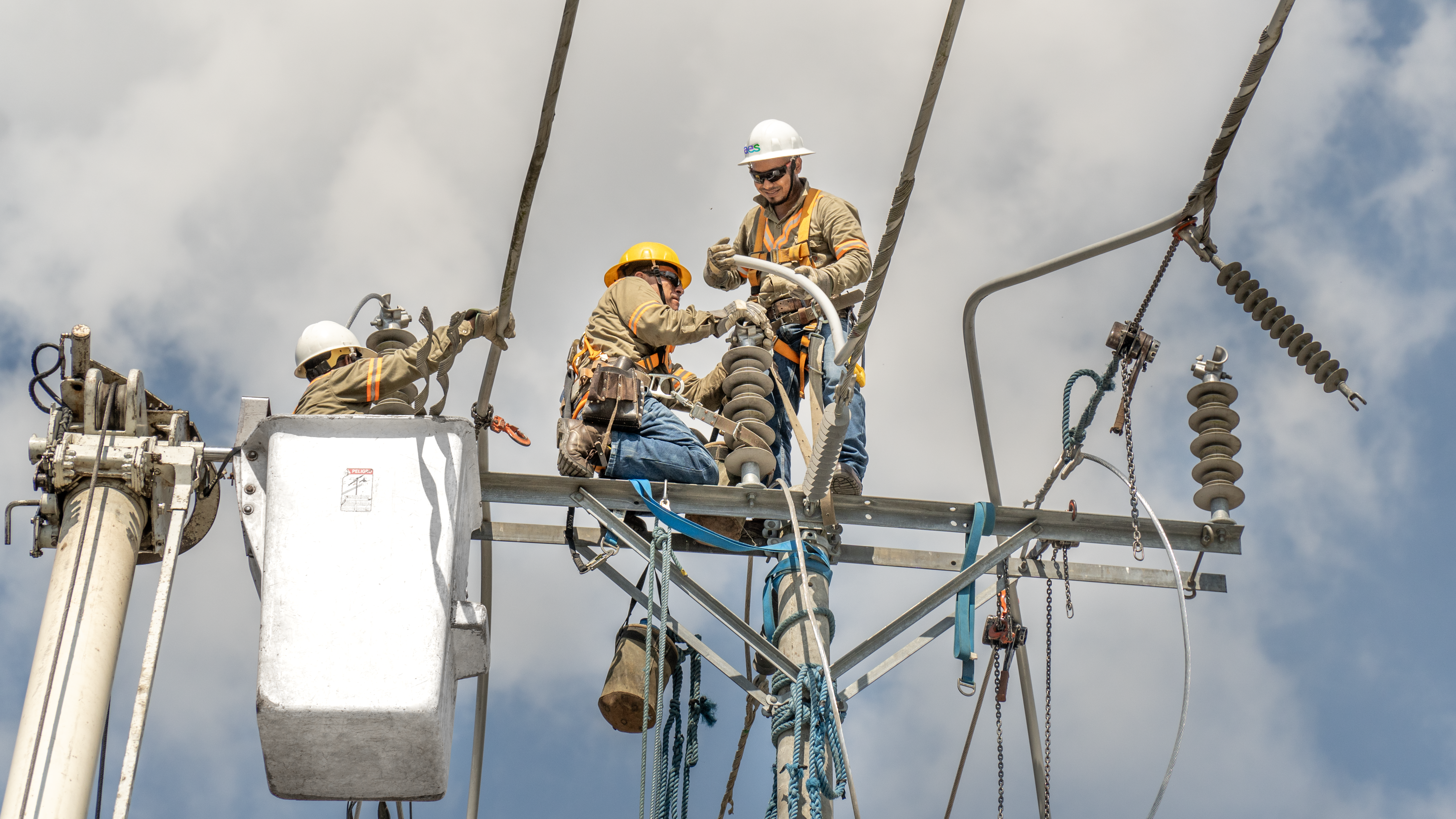 res trabajadores de líneas eléctricas con equipo de seguridad, incluidos cascos y arneses, están reparando un poste de electricidad. Dos de ellos están en el poste, uno de pie y otro sentado, manipulando componentes eléctricos.