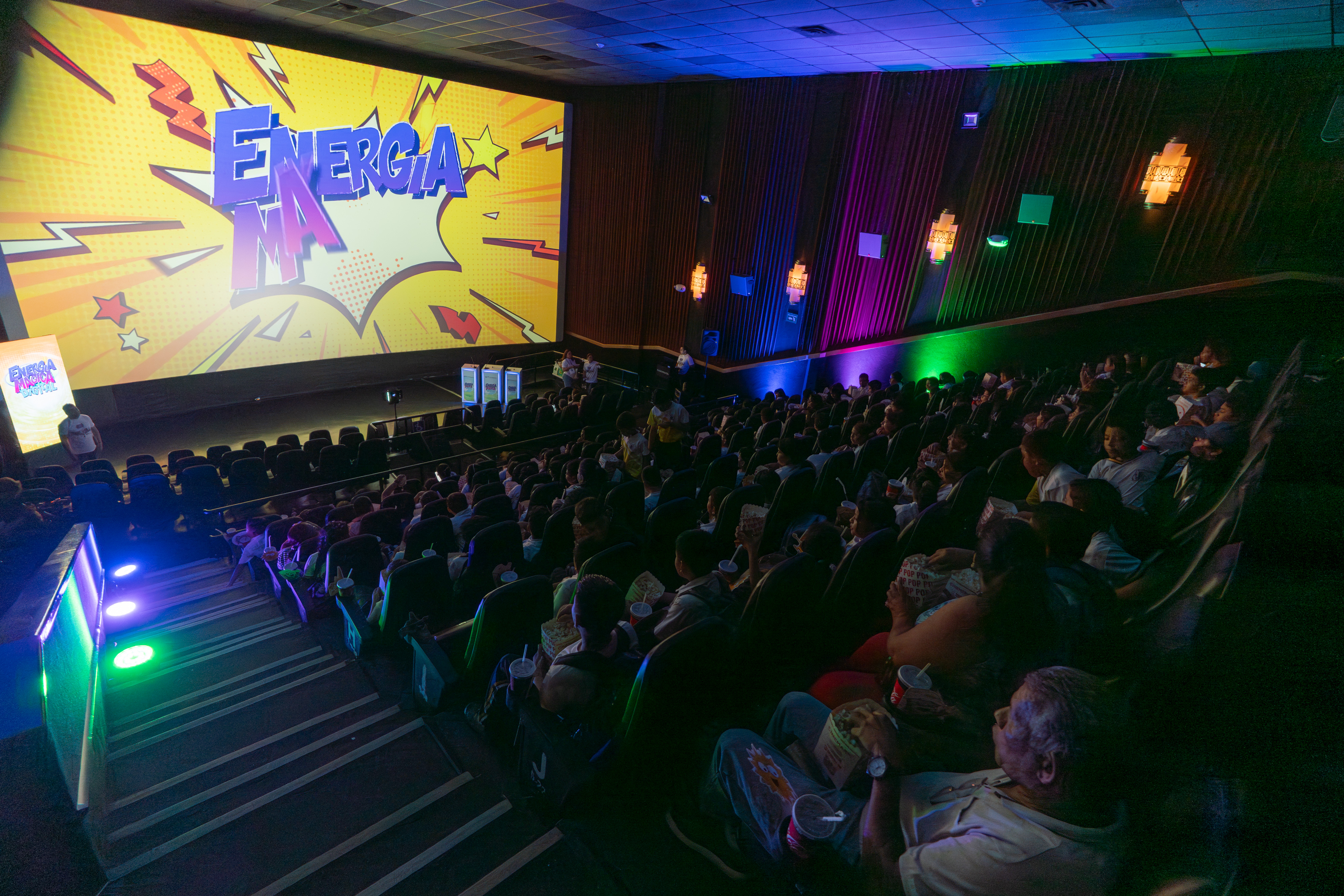Una vista amplia y ligeramente elevada muestra a una gran audiencia sentada en una sala de cine oscura. Todos miran hacia una pantalla de cine iluminada que muestra el texto "ENERGIA MAGICA DIGITAL" en un estilo de cómic con un fondo amarillo y elementos gráficos dinámicos.