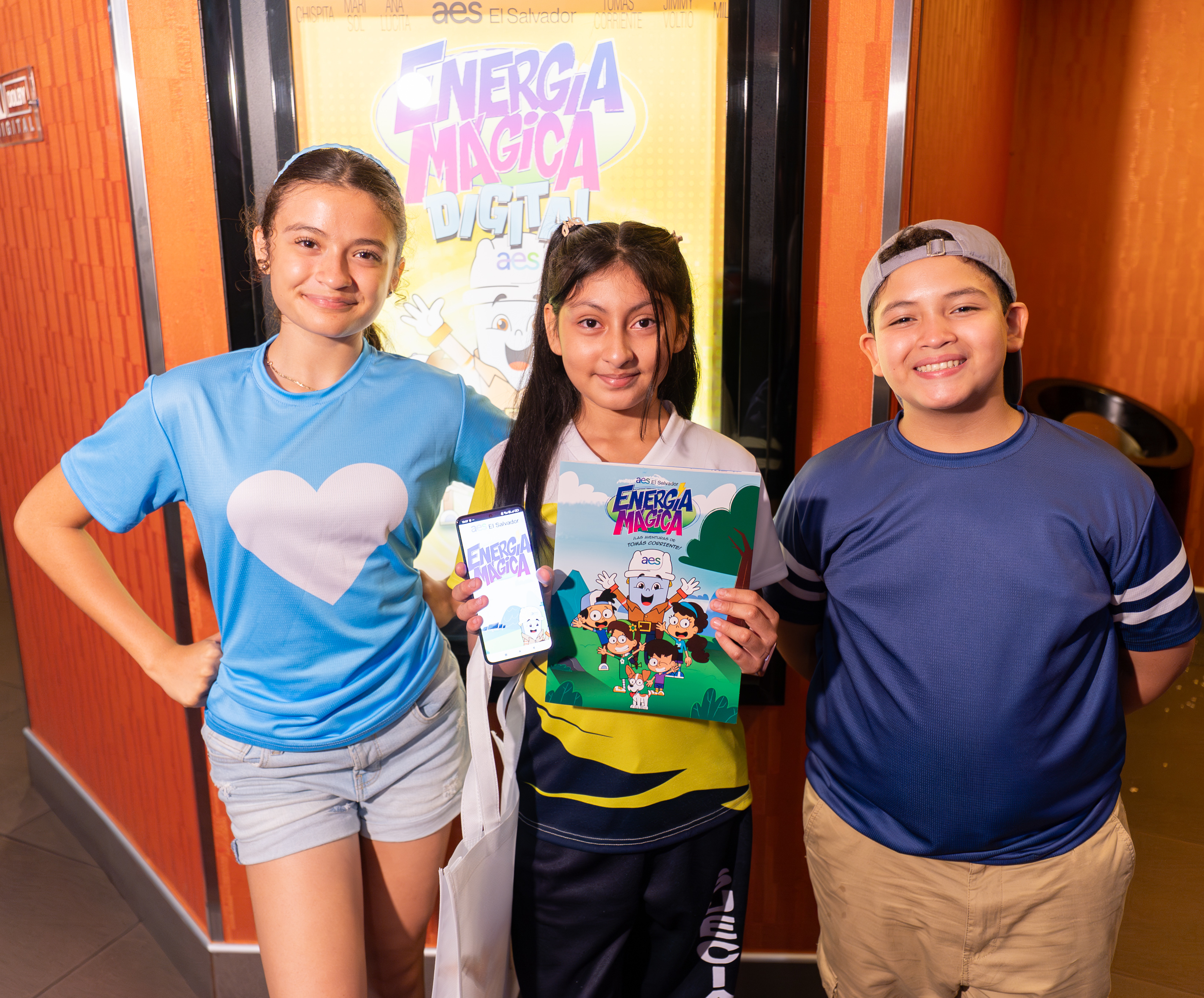 Tres niños sonrientes posan frente a un cartel de "Energia Magica Digital". La niña del centro sostiene una revista abierta del mismo título, mostrando dibujos animados de niños y un personaje parecido a un robot. La niña de la izquierda lleva una camiseta azul con un corazón blanco y sostiene un teléfono mostrando una aplicación de "Energia Magica". El niño de la derecha viste una camiseta azul oscuro y una gorra gris. El fondo muestra una pared texturizada de color naranja con un borde más oscuro a la izq