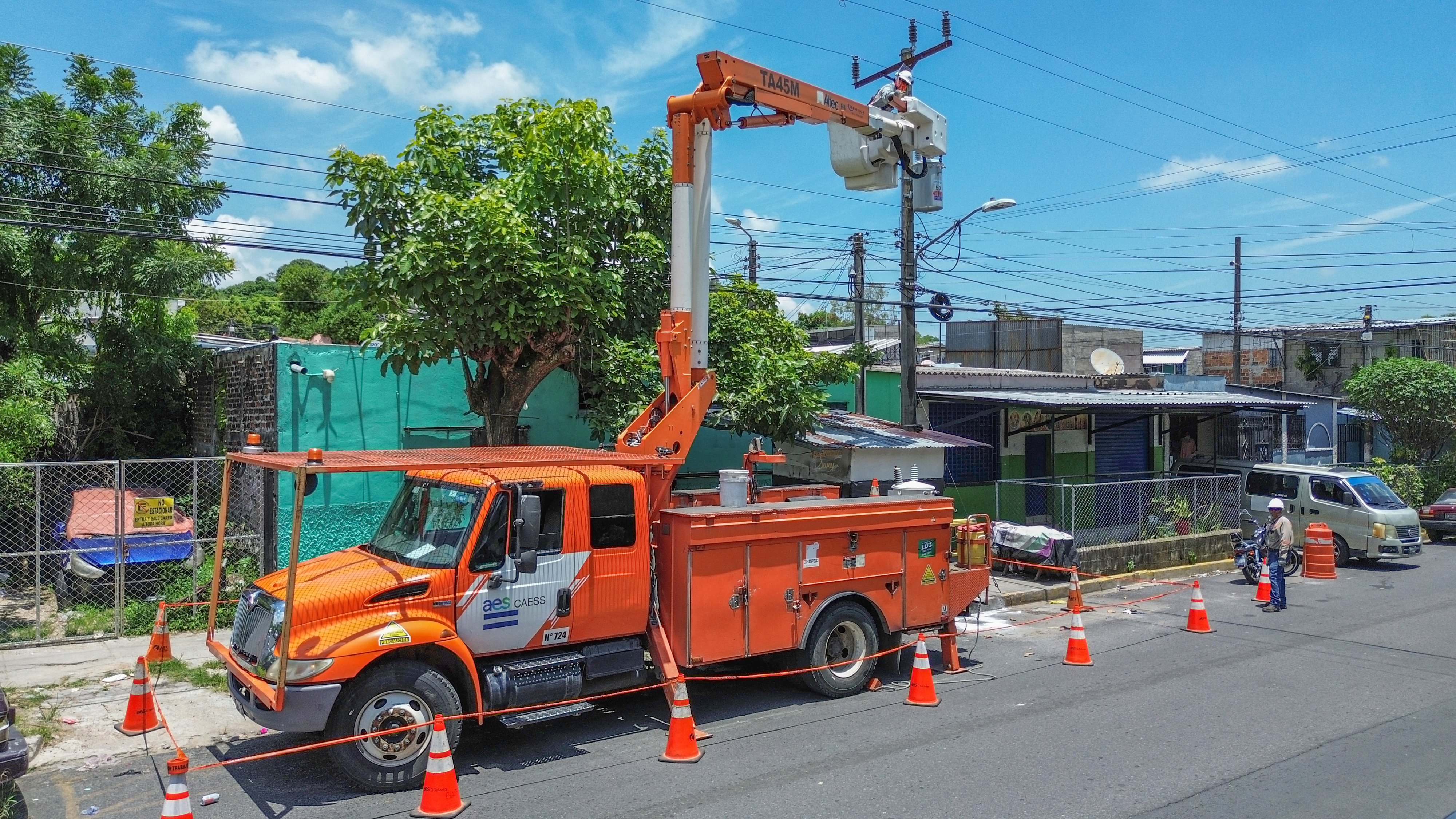 Un camión con pluma de mantenimiento eléctrico está estacionado en una calle bordeada de edificios bajos y vegetación. El camión, de color naranja, tiene las palabras "AES" y un número de serie. Hay conos de seguridad a su alrededor. En la pluma, un trabajador repara un cable. A la derecha, un segundo trabajador está de pie junto a una motocicleta.