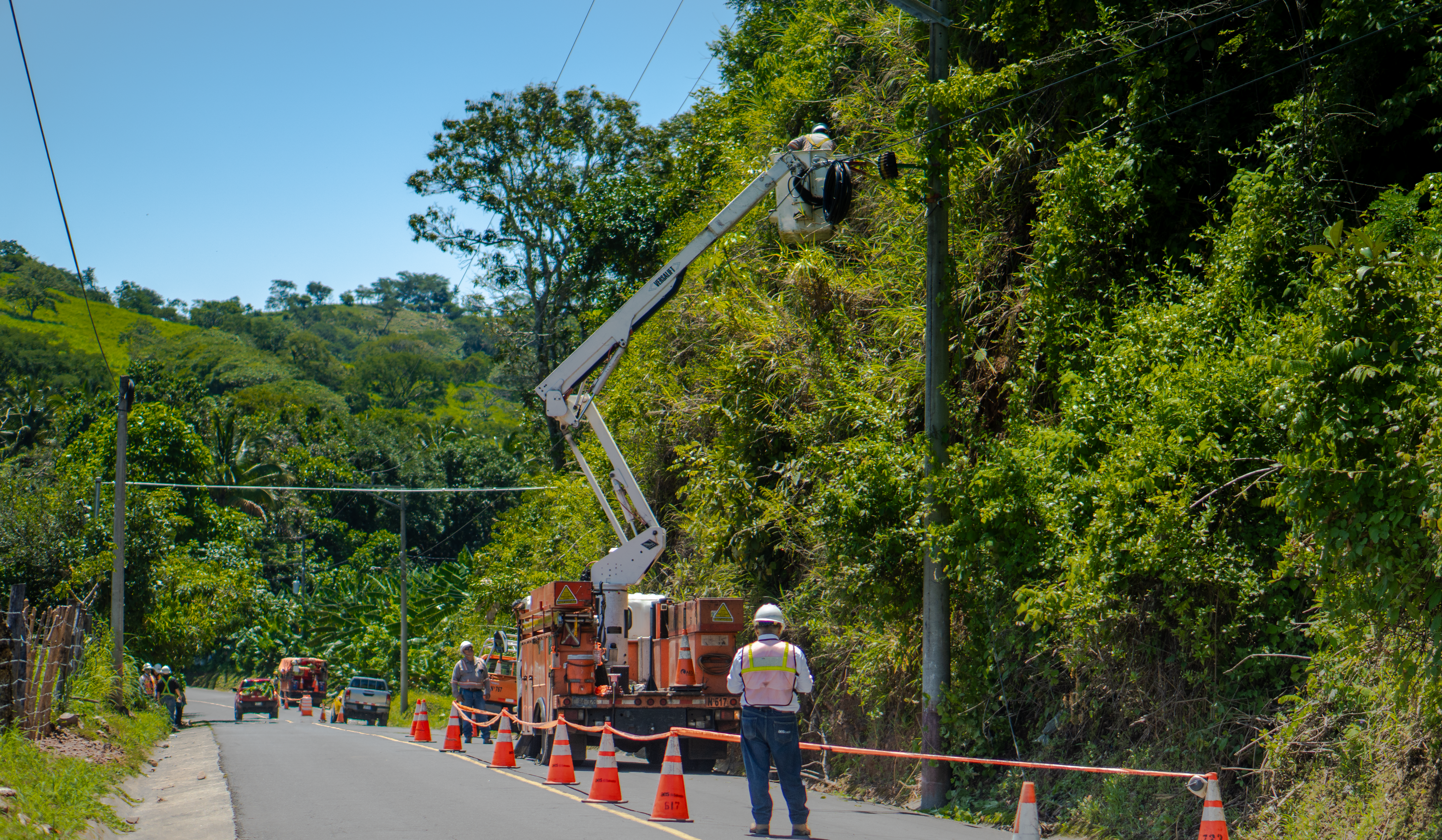 Un equipo de trabajadores eléctricos se encuentra en el lateral de una carretera rural. Un trabajador está en la canasta elevada de una grúa, reparando líneas en un poste de servicios públicos cubierto de vegetación. Varios compañeros están en el suelo, y el área de trabajo está señalizada con conos de tráfico y cinta naranja para seguridad. El fondo muestra colinas verdes y un cielo azul.