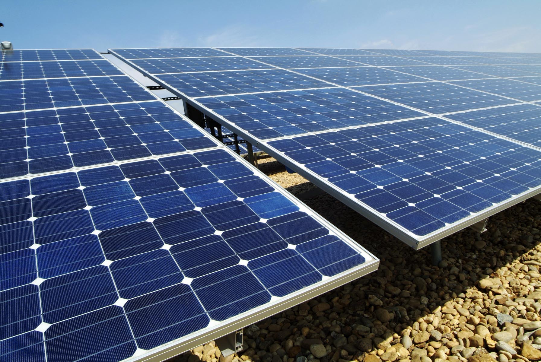 Primer plano de una serie de paneles solares azules y plateados dispuestos sobre una superficie de grava marrón. Los paneles reflejan la luz del sol bajo un cielo azul claro.