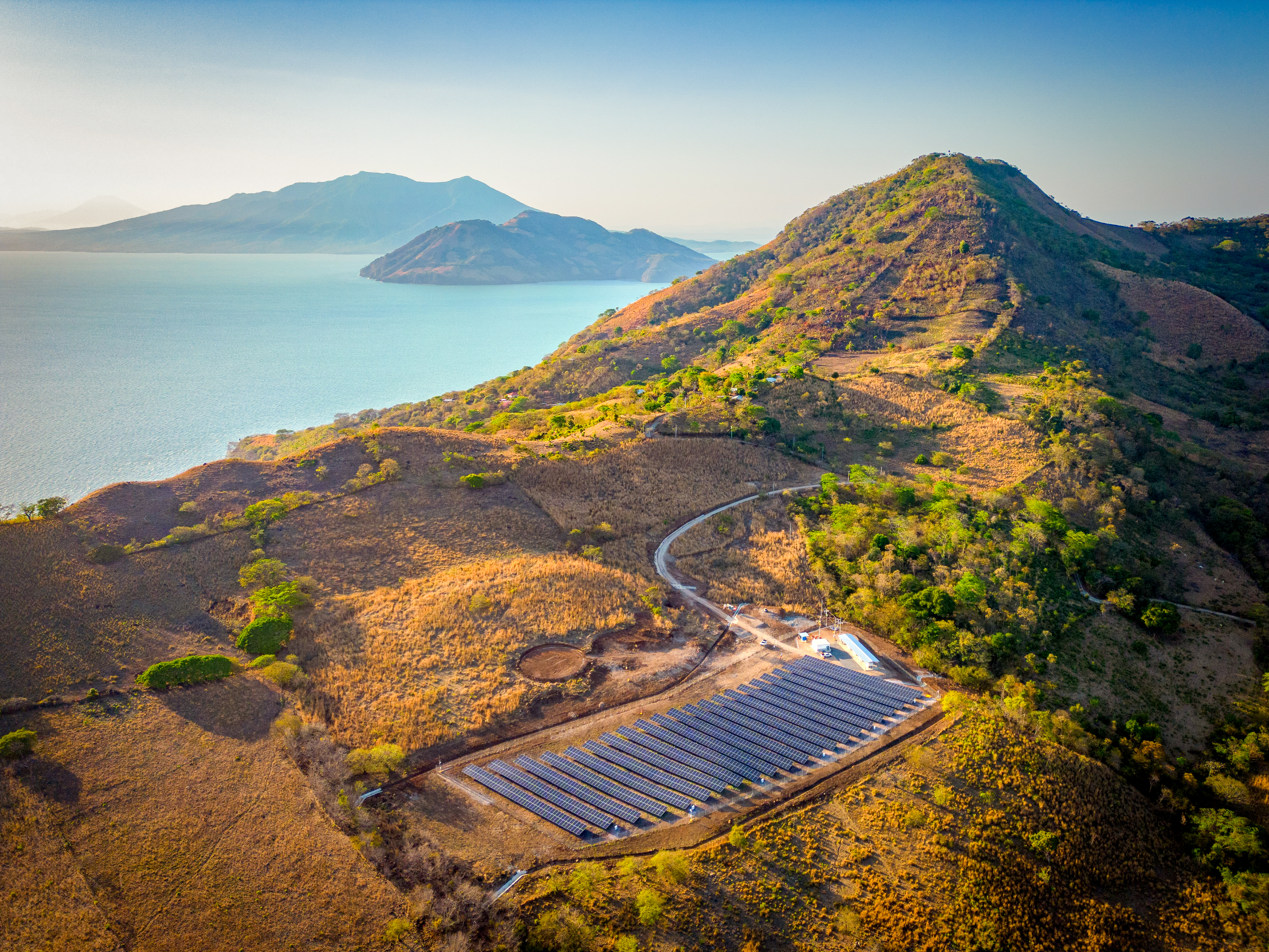 Vista aérea de un parque de paneles solares en una ladera con hierba seca. Un camino de tierra serpentea por las colinas. En el fondo, un gran cuerpo de agua refleja la luz, con islas montañosas que se alzan en la distancia. El cielo es claro y el sol ilumina la escena.