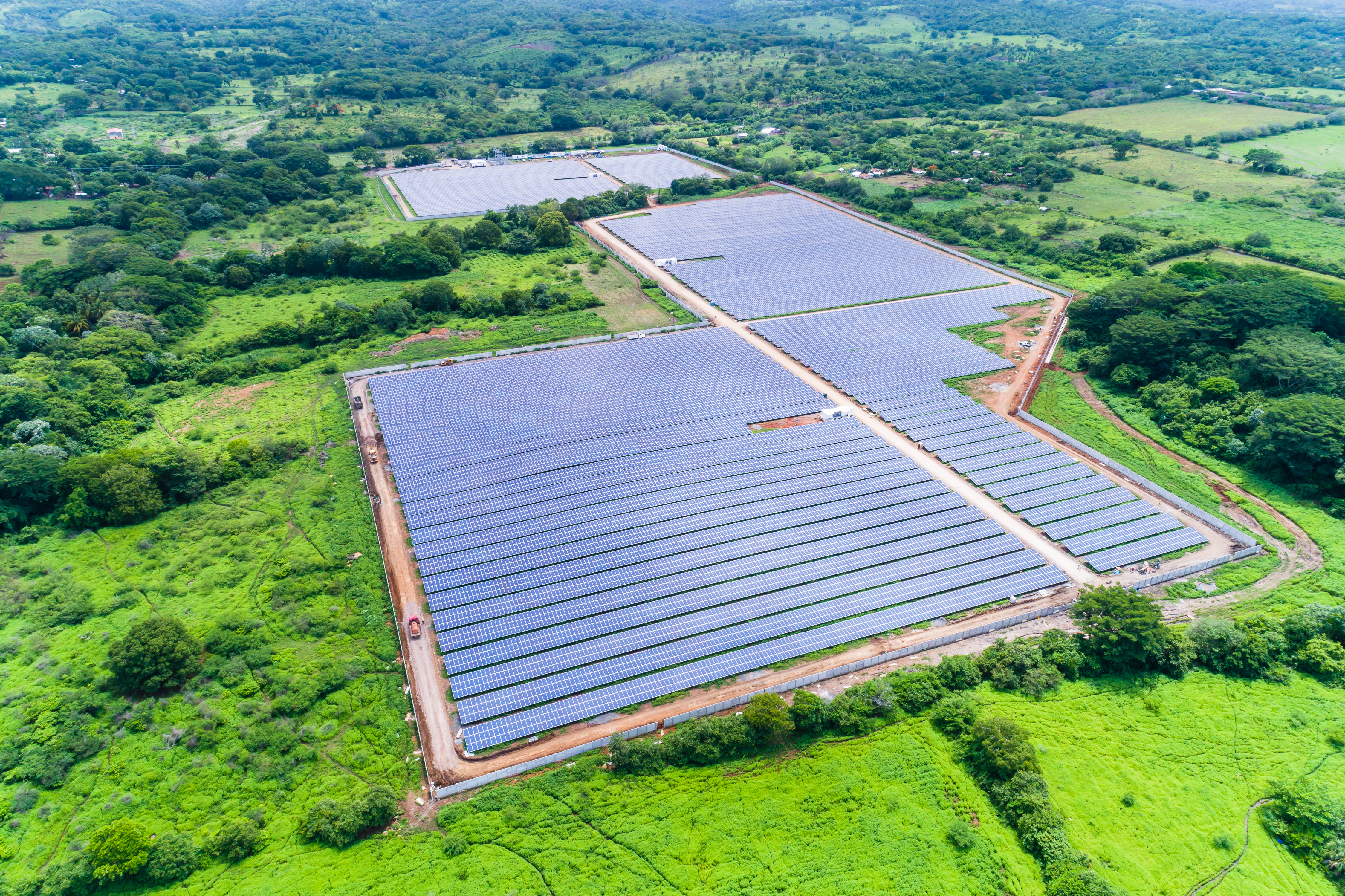 Vista aérea de una gran planta de energía solar, con múltiples campos de paneles fotovoltaicos dispuestos en largas filas. La planta está construida en una zona rural, rodeada de un paisaje vibrante de campos verdes y árboles. Caminos de tierra dividen las diferentes secciones de los paneles. El día es soleado.