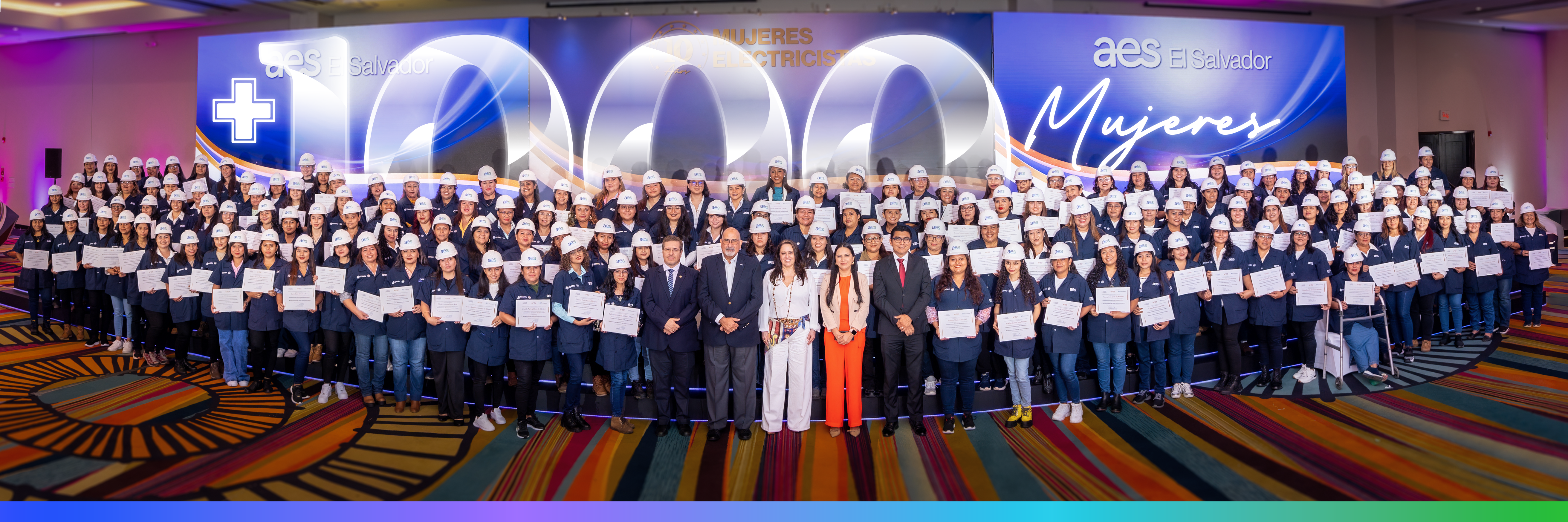 Un grupo grande de mujeres en uniformes de trabajo y cascos blancos, junto a algunos hombres con traje, posan frente a un escenario con el logo de "AES El Salvador" y el número "1000 Mujeres Electricistas". Las mujeres sostienen diplomas y certificados.