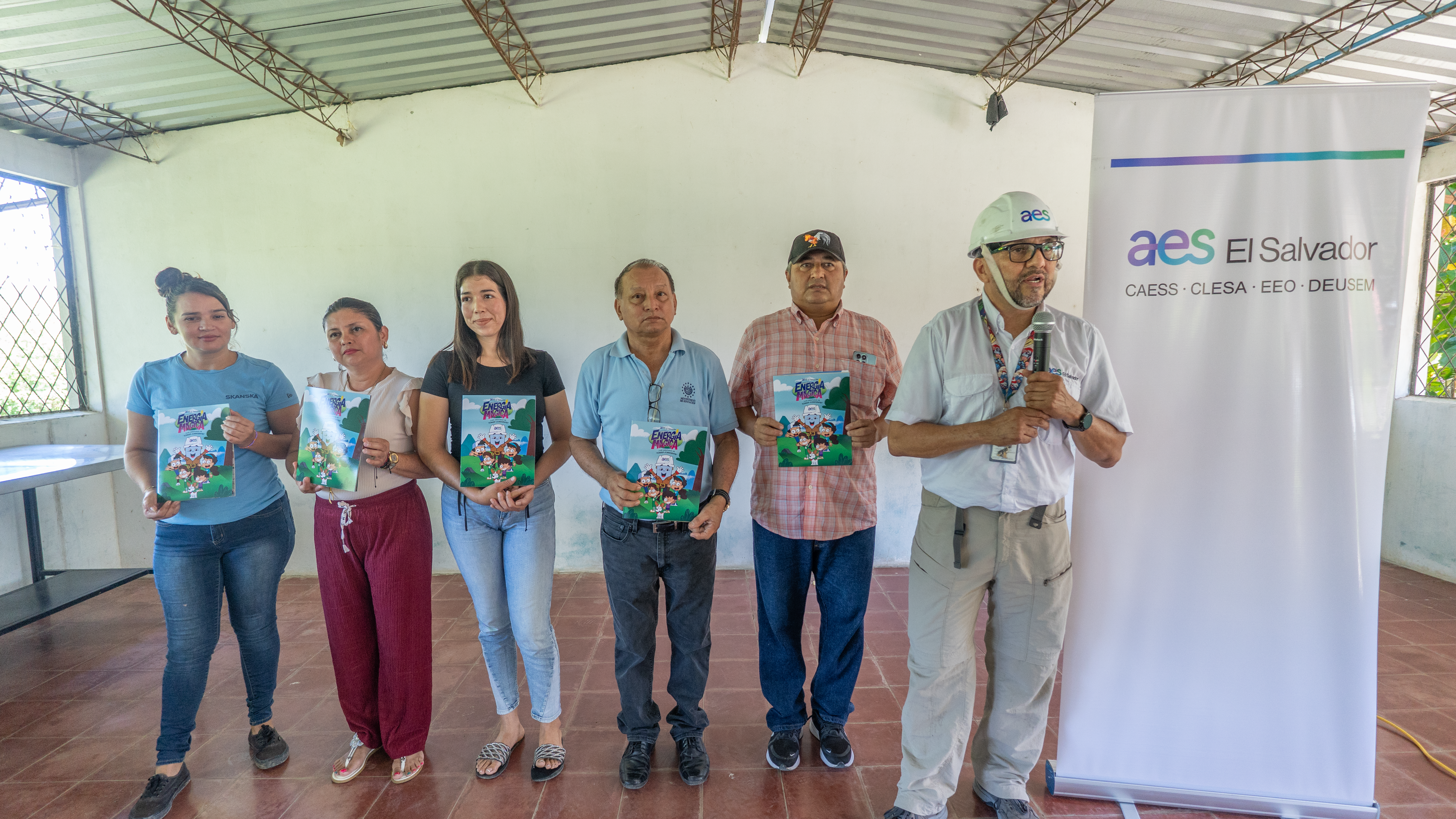 Seis personas posan para una foto de grupo en una sala de presentaciones. Un hombre de mediana edad con un casco blanco y un micrófono sostiene folletos, mientras que un grupo de cinco personas a su lado, de edades variadas, sostiene los mismos folletos. Detrás de ellos hay un cartel de la empresa AES El Salvador.