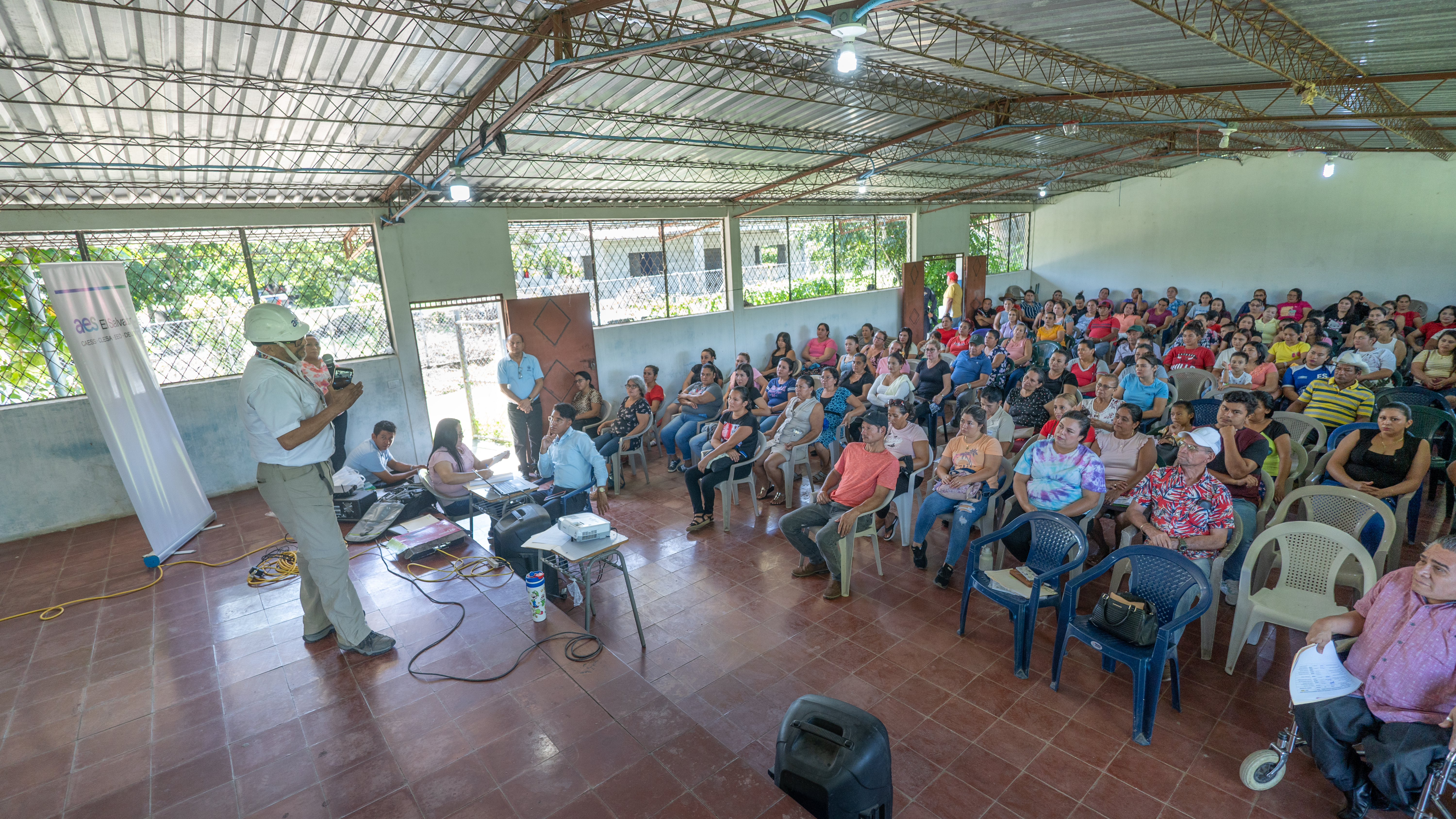 Una vista de interior de una sala de presentación. En el lado izquierdo, un hombre blanco con un chaleco de alta visibilidad, casco, camisa de manga larga blanca y pantalones caqui está de pie, hablando a una gran audiencia. Hay un proyector, una pantalla, y un estandarte detrás de él. En el lado derecho y en el centro hay un grupo diverso de hombres y mujeres sentados en sillas de plástico.