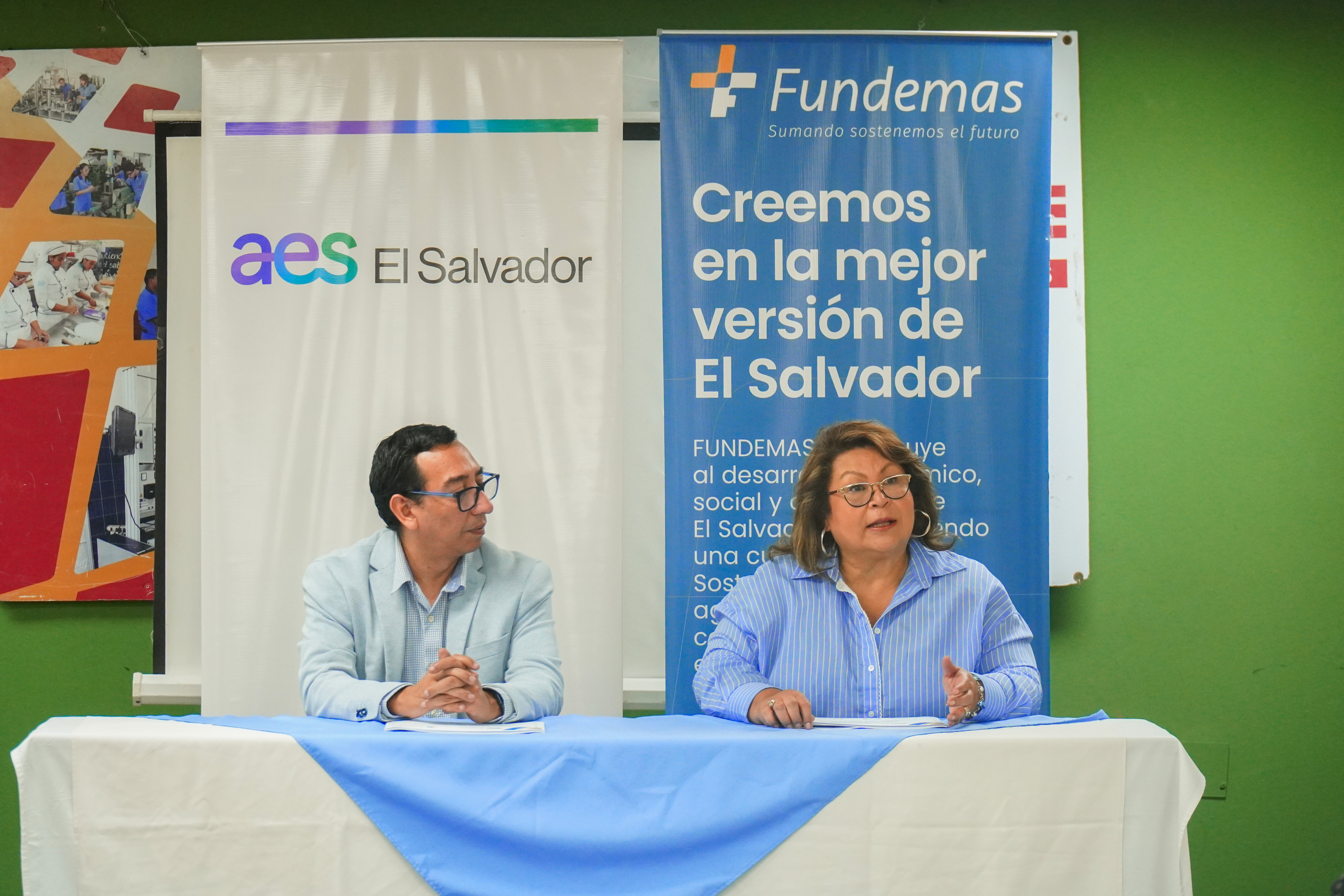 Dos presentadores, un hombre y una mujer, se sientan detrás de una mesa cubierta con manteles blancos y azules. Detrás de ellos hay dos pancartas grandes. La de la izquierda es blanca con el logotipo de "aes El Salvador" en púrpura. La de la derecha es azul con el logotipo de "Fundemas" en la parte superior y el texto "Creemos en la mejor versión de El Salvador" en el centro, con texto adicional debajo.