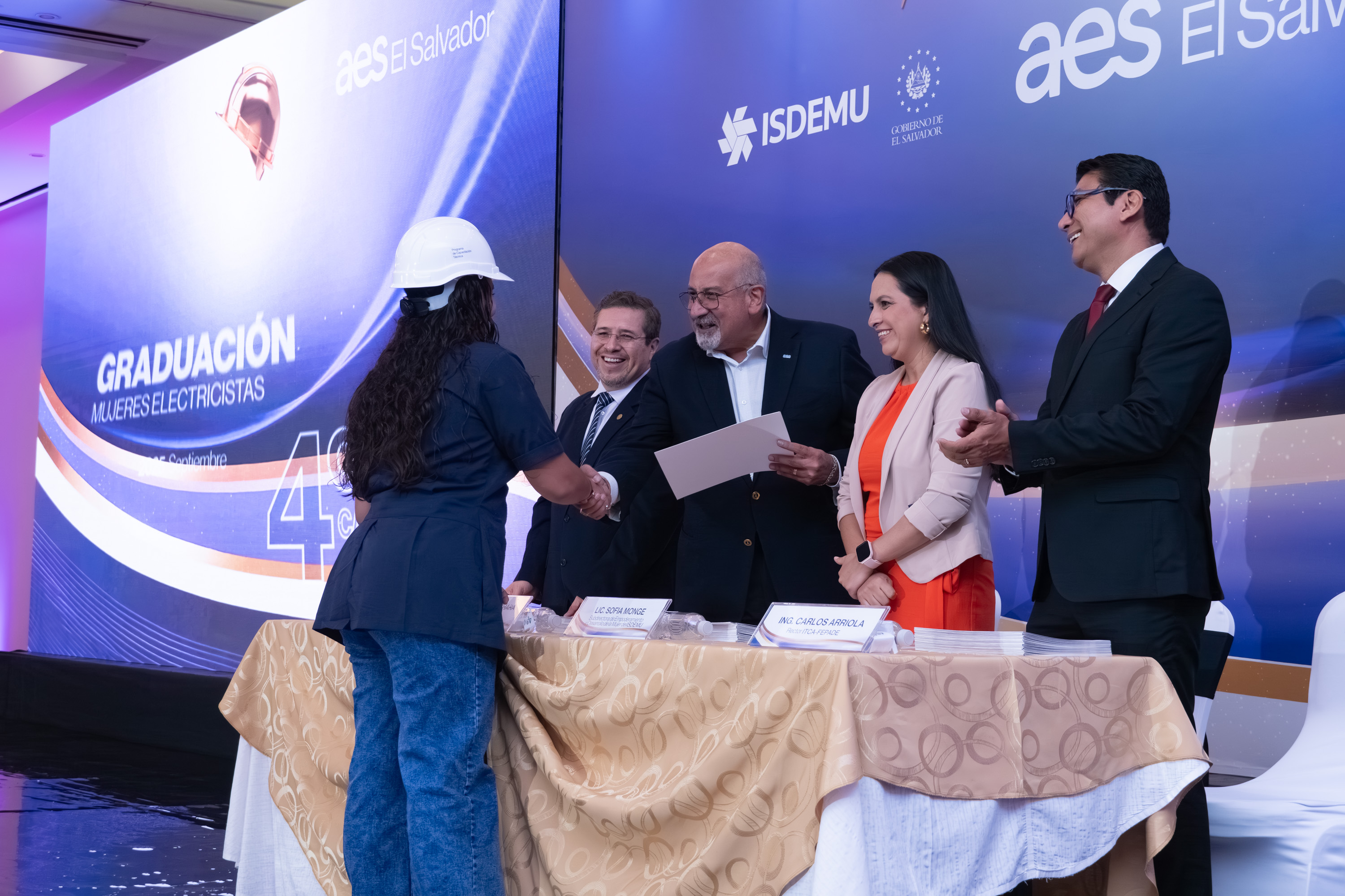 Una mujer, vestida con uniforme de trabajo azul, jeans y casco blanco, estrecha la mano de un hombre mientras recibe un certificado en un escenario. Detrás de una mesa con un mantel dorado, otros tres individuos (dos hombres y una mujer) sonríen. El telón de fondo del escenario muestra "GRADUACIÓN MUJERES ELECTRICISTAS" y logotipos de "AES El Salvador".