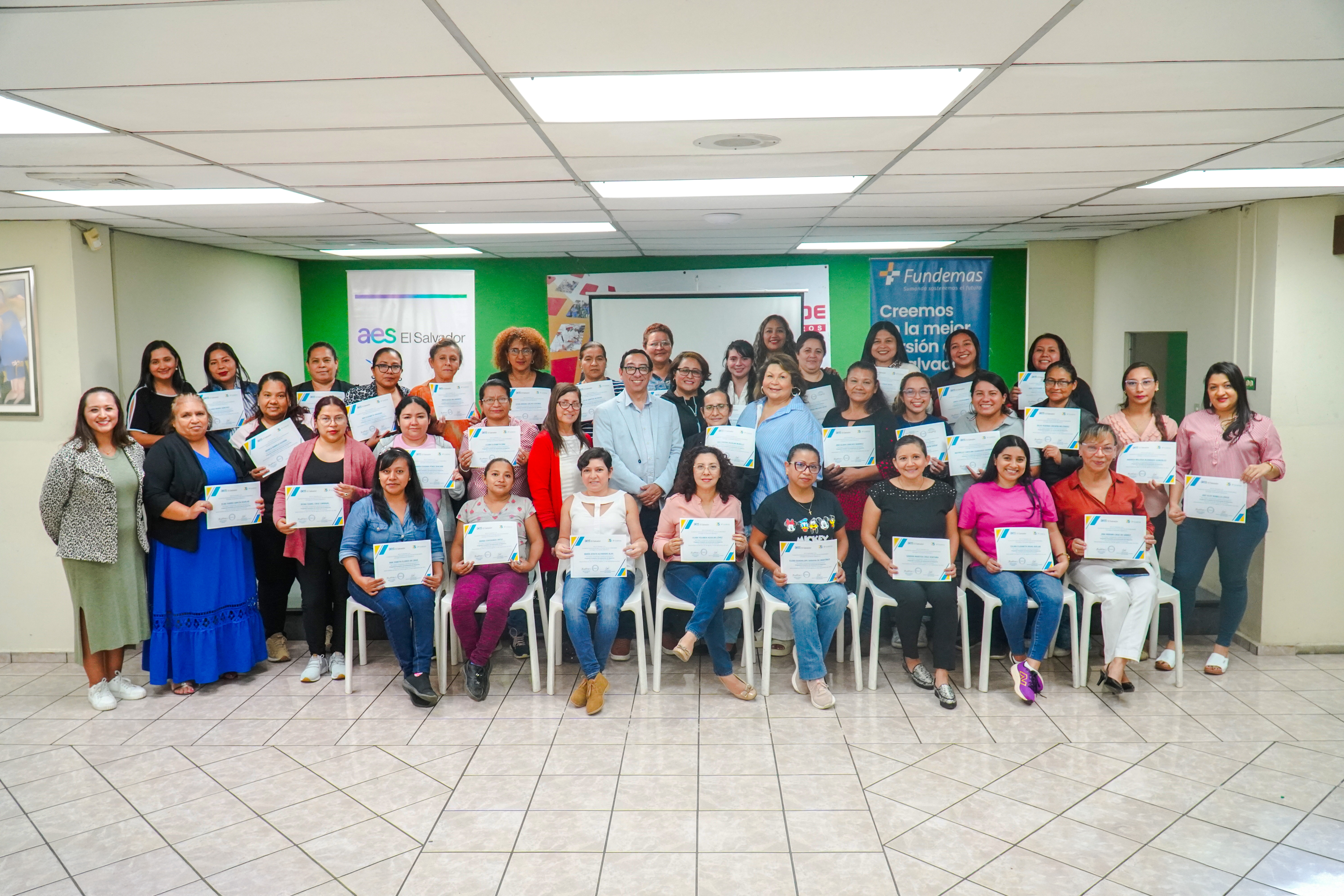 Un grupo diverso de unos treinta hombres y mujeres, en su mayoría mujeres, sonríen mientras posan en una oficina bien iluminada. La mayoría sostienen diplomas o certificados. Detrás de ellos hay una pared de color verde brillante con los logotipos de "AES El Salvador" y "Fundemas", con el lema "Creemos en la mejor inversión, la inversión social".