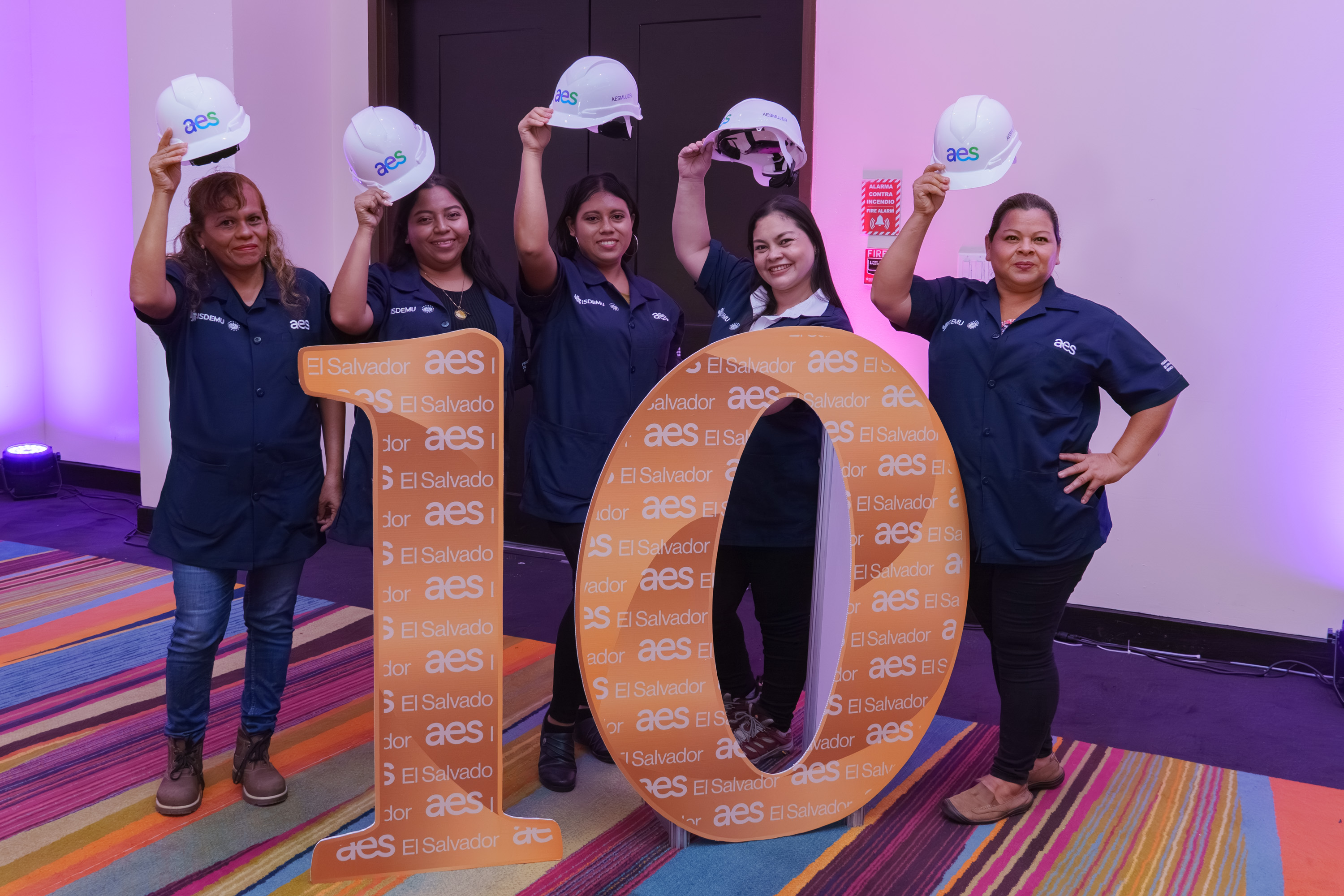 Cinco mujeres, vestidas con uniformes de trabajo azules, sostienen sus cascos blancos de "AES" en alto mientras posan alrededor de un gran número "10" de cartón, decorado con el logo "AES El Salvador". Las mujeres sonríen, celebrando los 10 años de un programa o iniciativa, probablemente "Mujeres Electricistas".